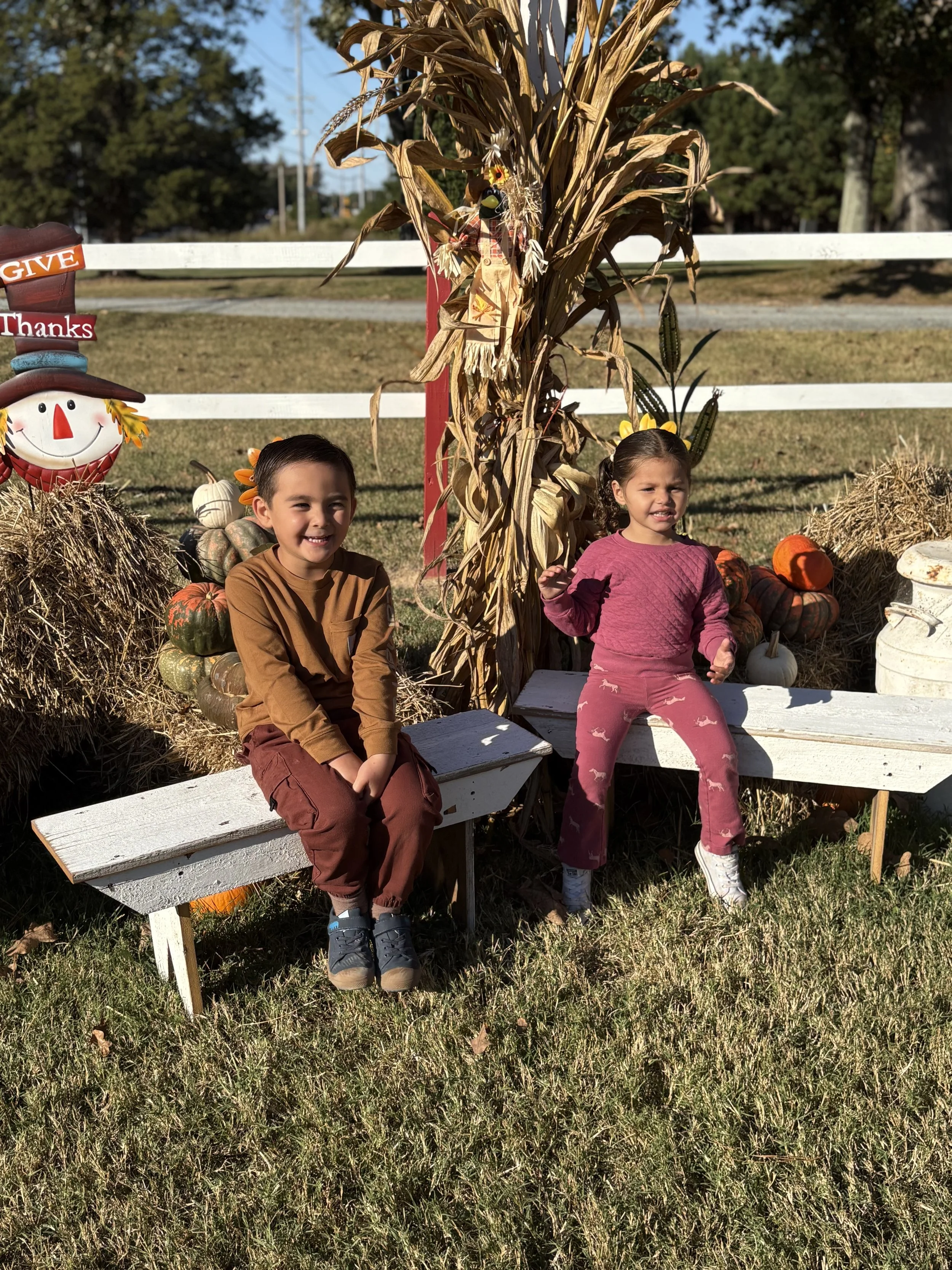 Two children sitting on white weathered benches outdoors in front of a fall-themed display with hay, pumpkins, and a decorative scarecrow made of dried corn stalks. A smiling boy is on the left wearing a brown long-sleeved shirt and matching pants, while a girl on the right wears a pink sweater and pink pants with white boots.