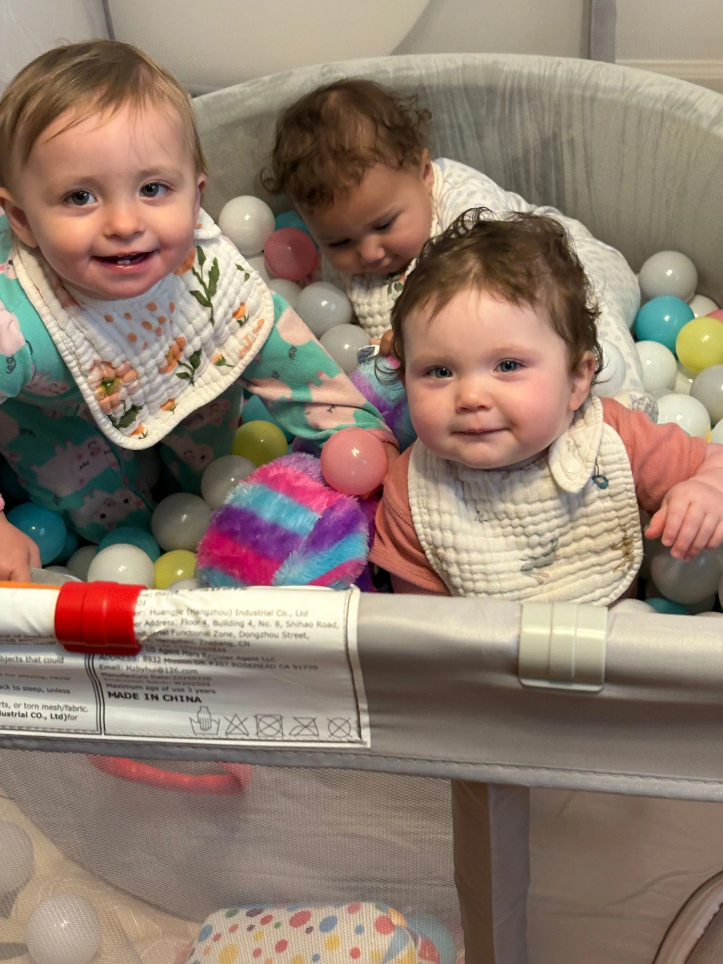 Three young children playing in a ball pit filled with colorful plastic balls. One child is smiling at the camera, while the other two are focused on playing.