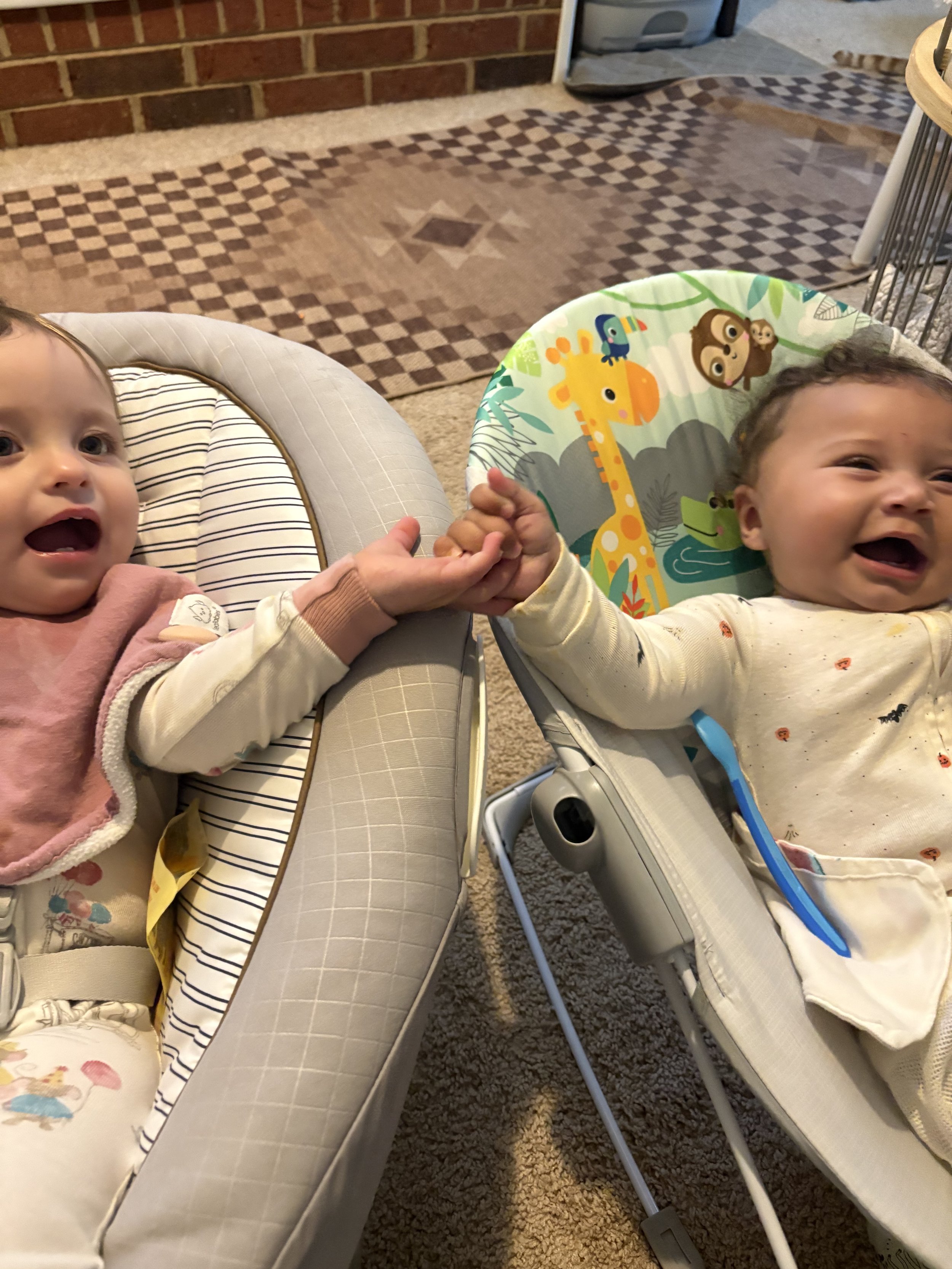Two babies in high chairs holding hands and smiling, with one baby wearing a pink jacket and the other wearing a cream-colored shirt with small orange and black patterns. Behind them is a patterned area rug.