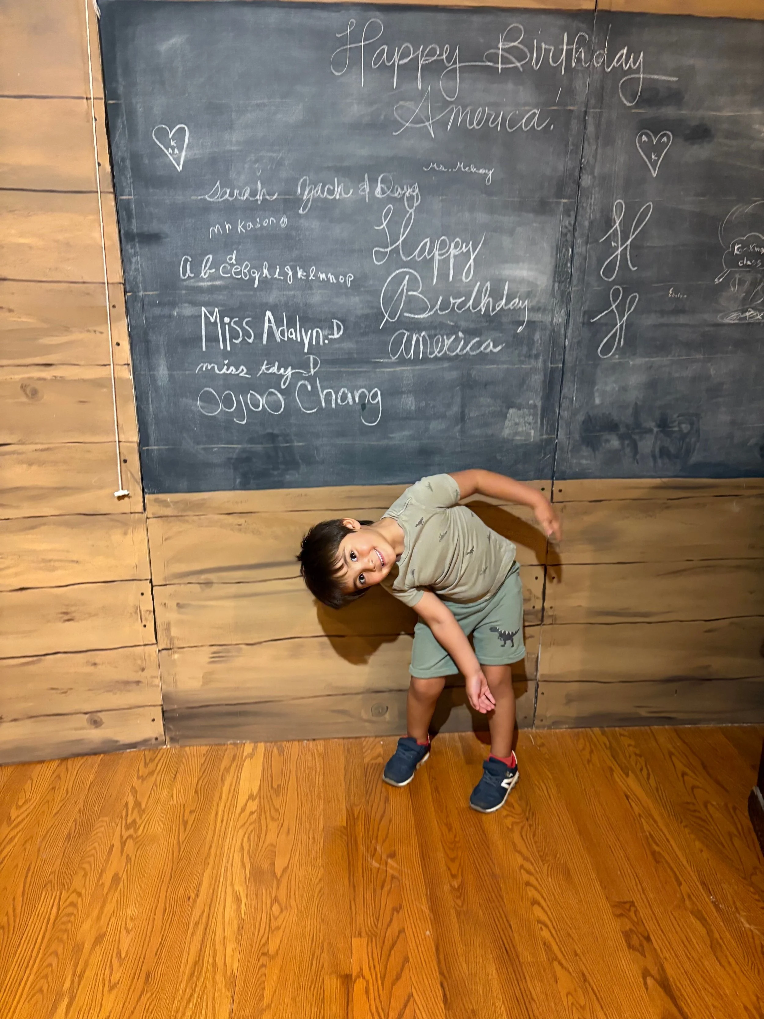 Grinning young boy in green shorts and gray t-shirt leans to the side and stands in front of a wooden wall and chalkboard with birthday messages and signatures for America.