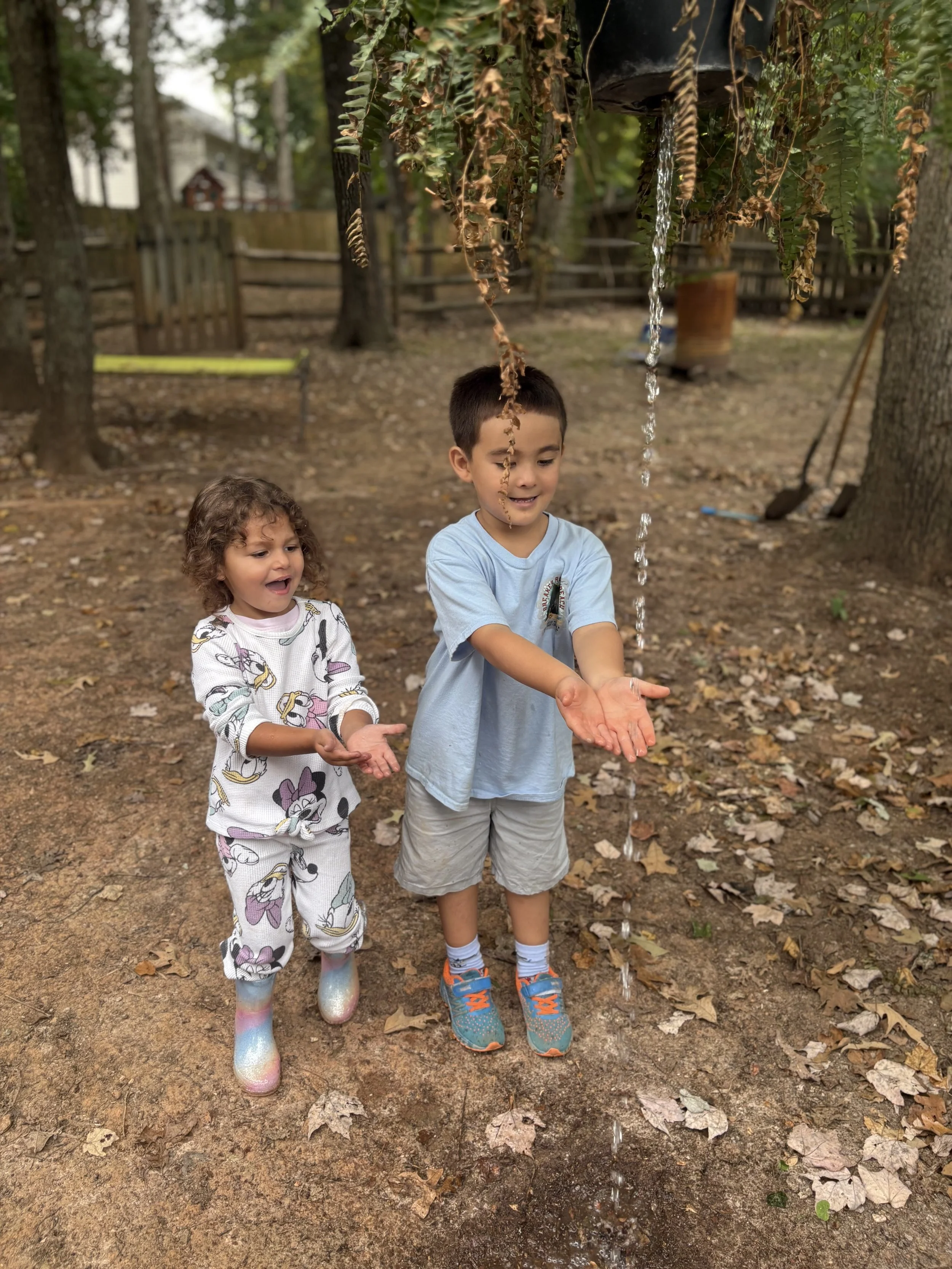 Two children, a girl and a boy, playing with water under a hanging spout outdoors in a wooded area. The girl is wearing pajamas with Disney characters and rain boots, and the boy is wearing a light blue T-shirt and shorts with sneakers. They appear happy and engaged with the water.