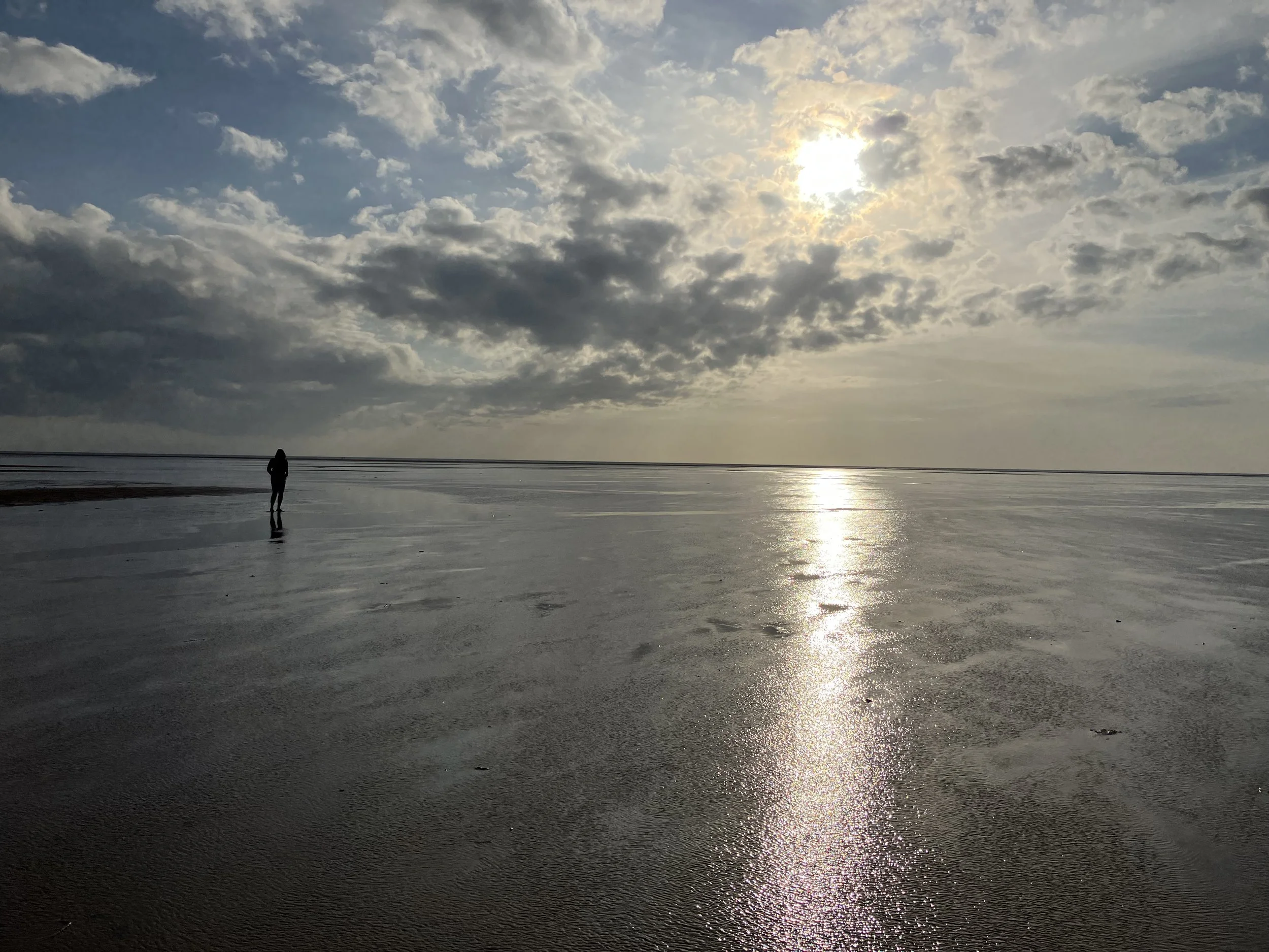 A person standing on a reflective wet beach during sunset with a partly cloudy sky.