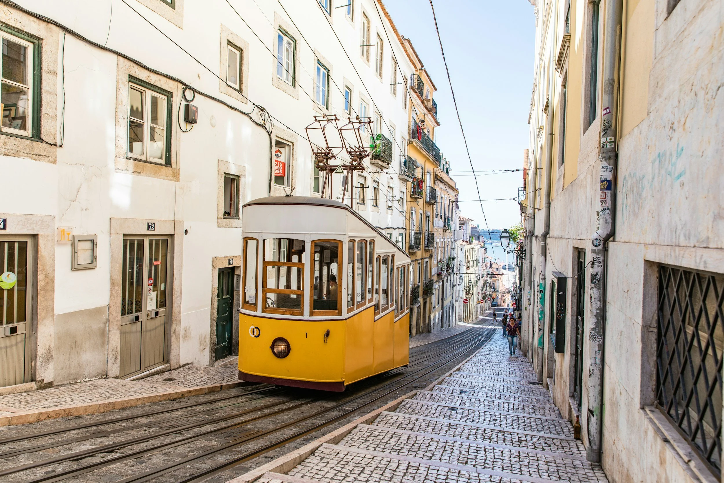 Iconic yellow Bica tram traveling up a steep, cobblestone street in Lisbon, Portugal, lined with traditional whitewashed buildings and balconies, capturing the city’s historic charm and vibrant urban life.