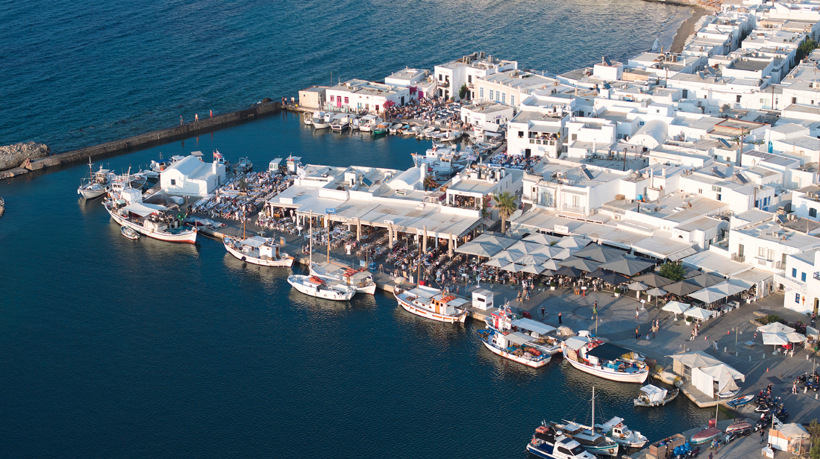Aerial view of the whitewashed harbor and waterfront restaurants in Paros, Greece, near Andronis Minois hotel, with boats docked along the Aegean Sea.