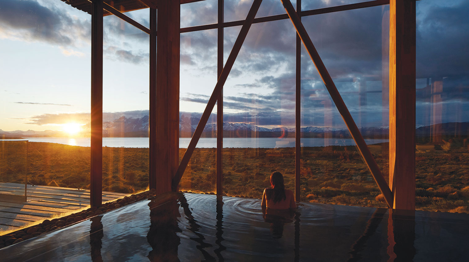Woman relaxing in heated spa pool with floor-to-ceiling windows framing Patagonian mountains and lake at sunset in Chile