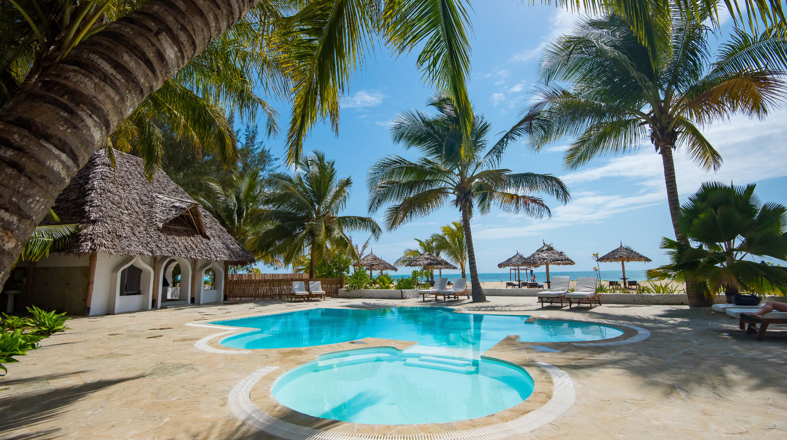 Amani Boutique Hotel in Zanzibar, Tanzania, featuring a tropical pool surrounded by palm trees, thatched-roof cabanas, and direct access to a white sand beach and turquoise Indian Ocean.