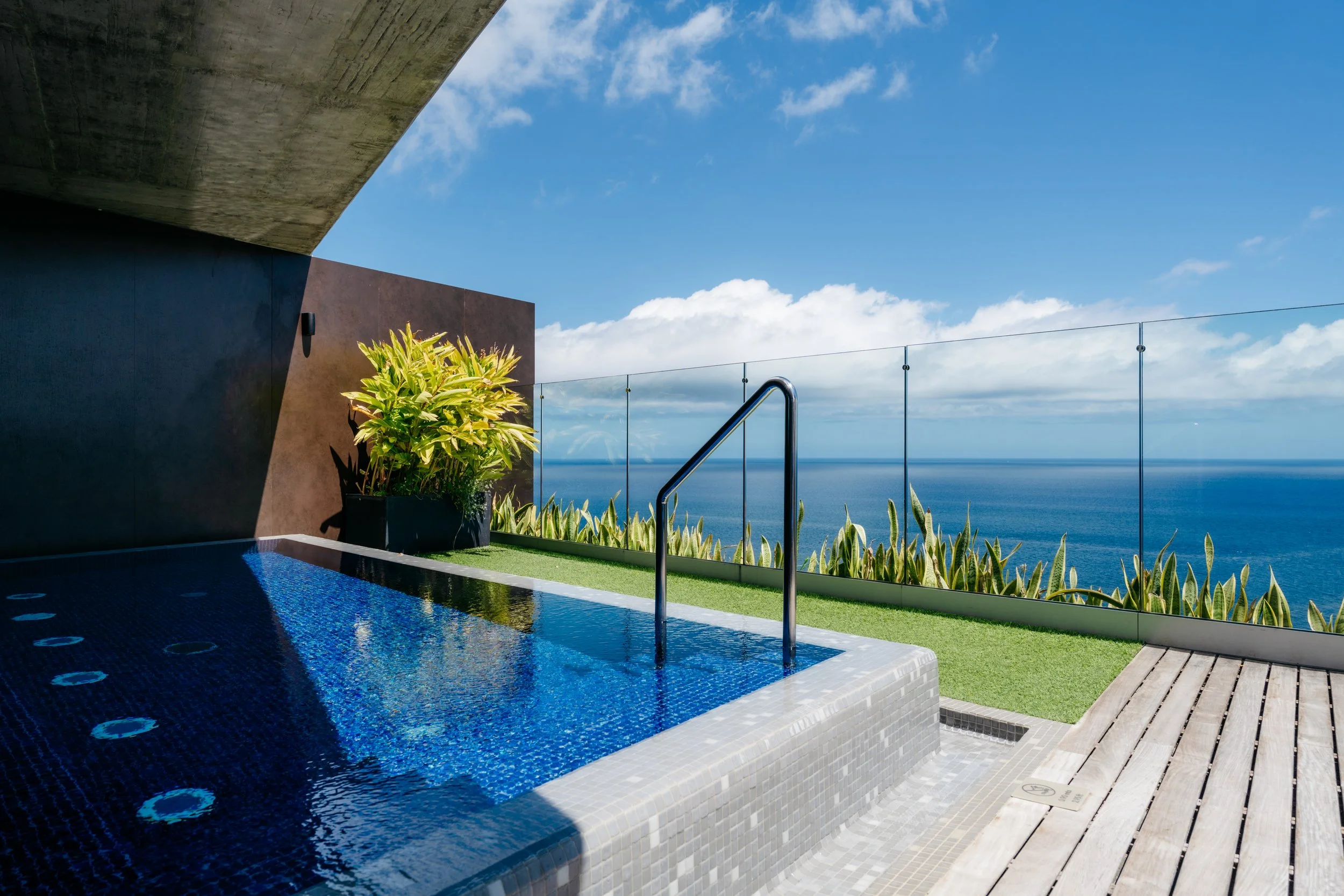 Modern private plunge pool with glass railing, tropical plants, and panoramic Atlantic Ocean views—clean lines, minimalist design, and natural light in Funchal, Madeira.