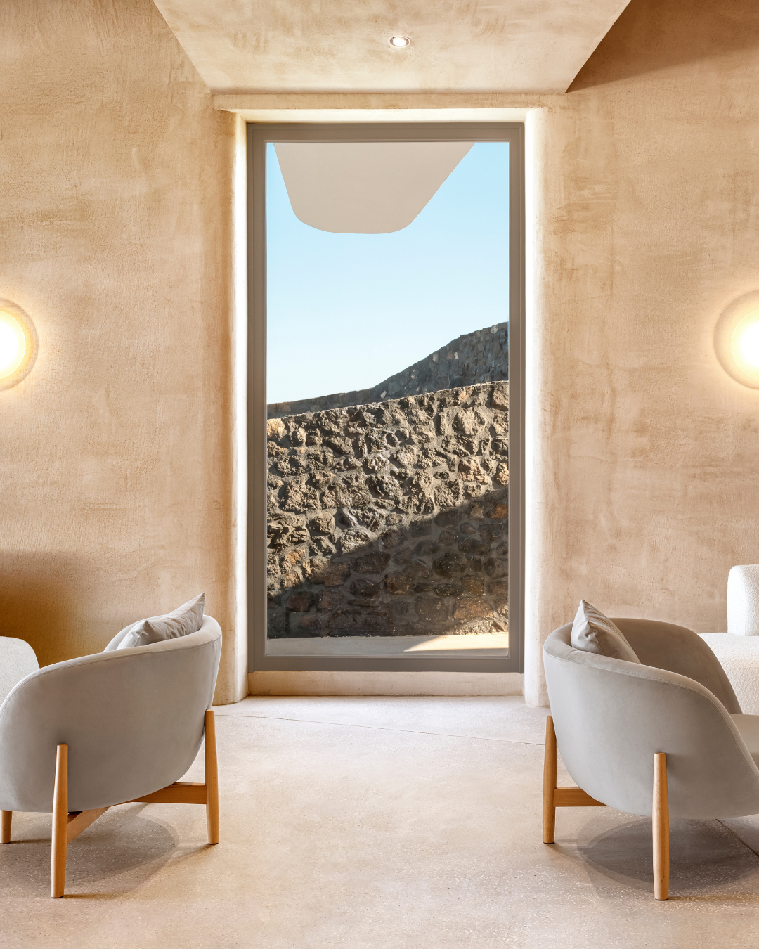 Interior of a modern room with a large floor-to-ceiling window showing a stone wall and clear sky outside. The room features beige textured walls, two white armchairs with cushions, and soft ambient lighting.
