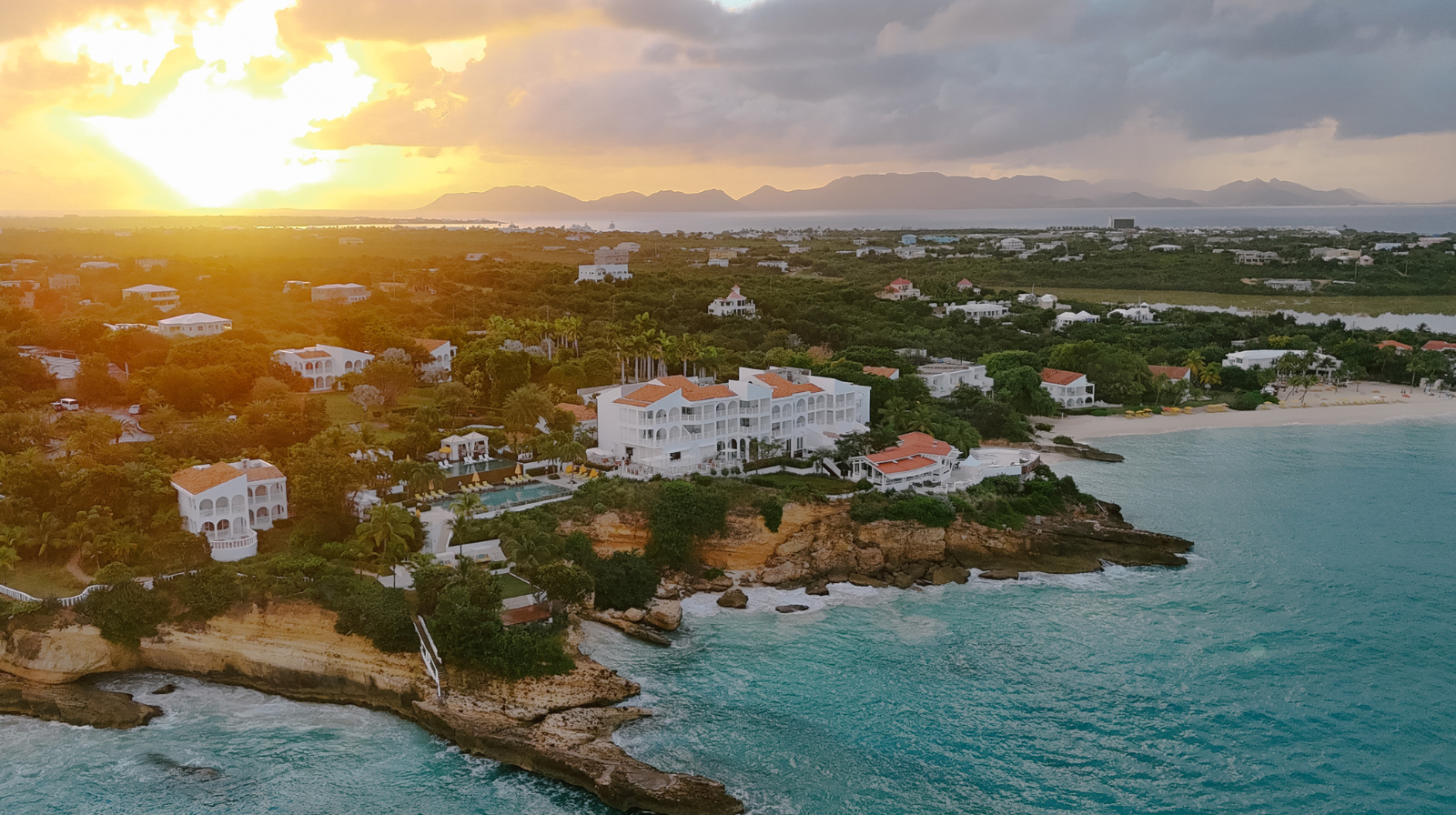 Aerial view of Malliouhana Resort in Anguilla at sunset, showcasing luxury oceanfront accommodations, white villas, turquoise waters, and lush island landscape.