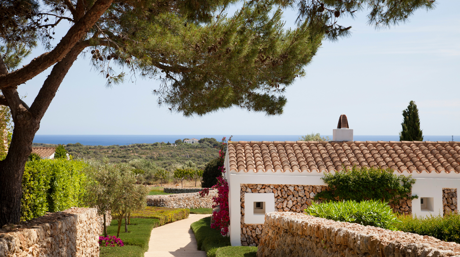Pathway through Mediterranean gardens at Torralbenc boutique hotel in Menorca, Spain, with traditional stone architecture and sea views in the distance.