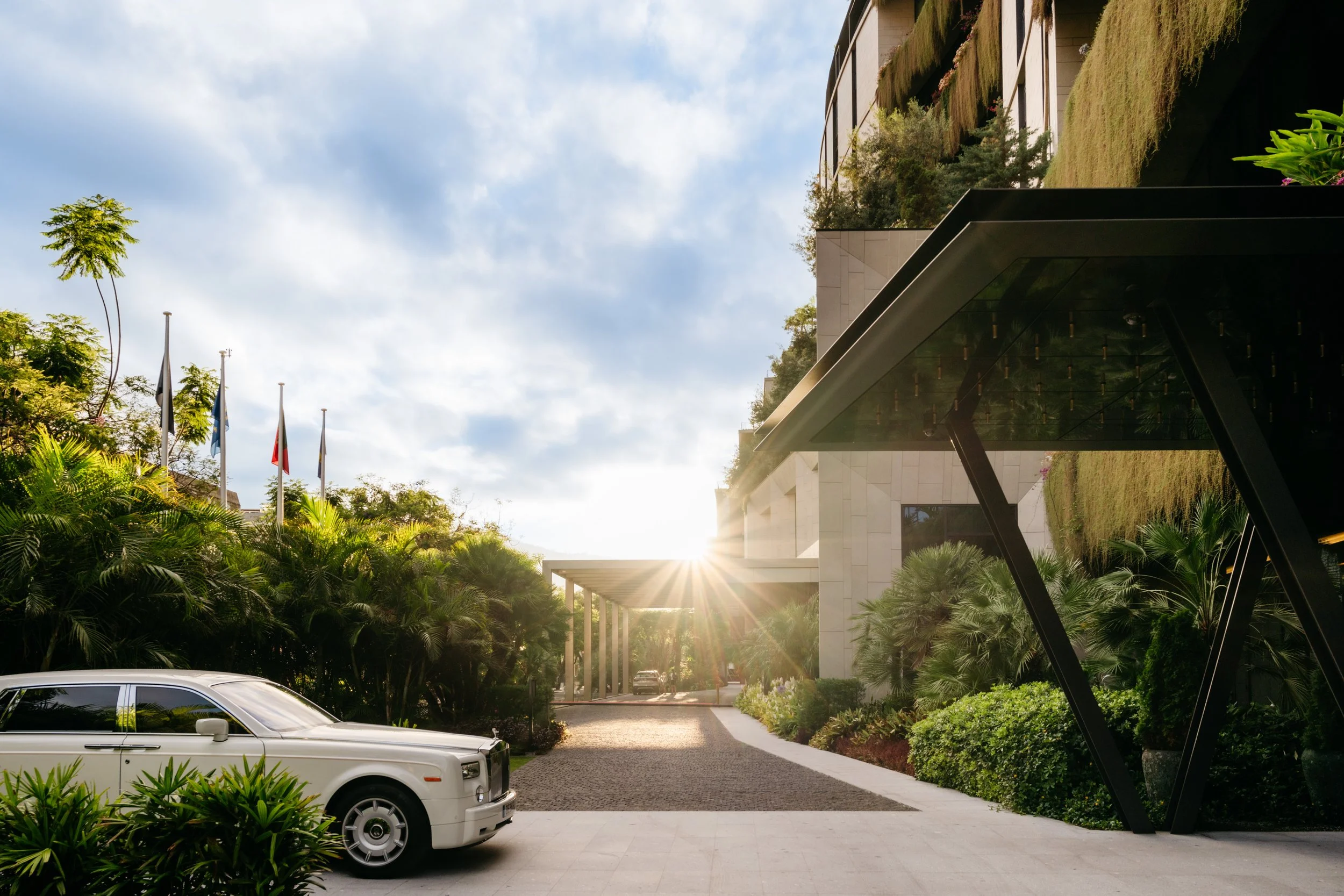 Modern luxury hotel entrance with lush tropical landscaping, a vintage white Rolls-Royce, and dramatic architectural lines at sunrise—greenery and understated opulence.