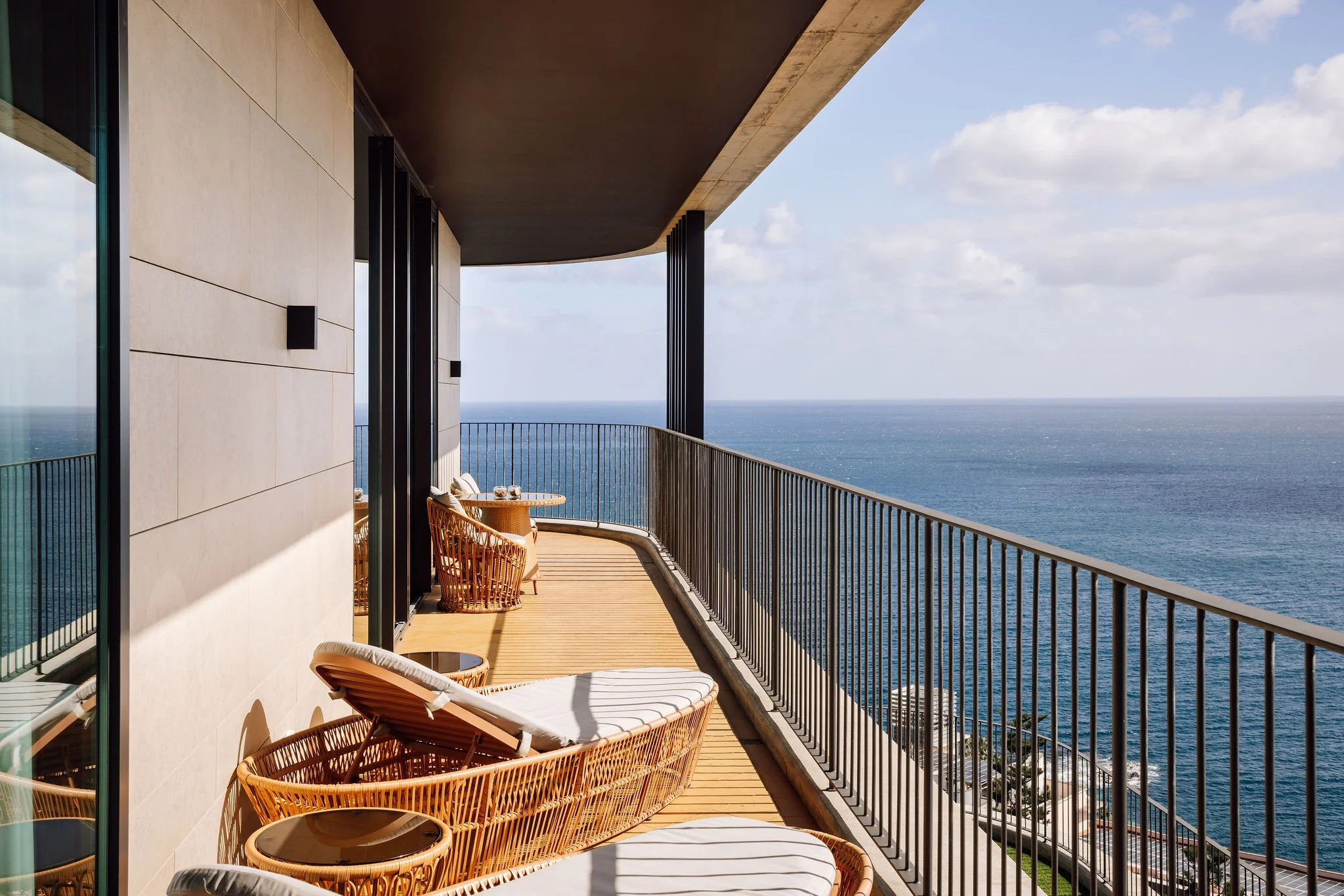 Balcony with rattan lounge chairs and small tables overlooking the ocean under partly cloudy sky