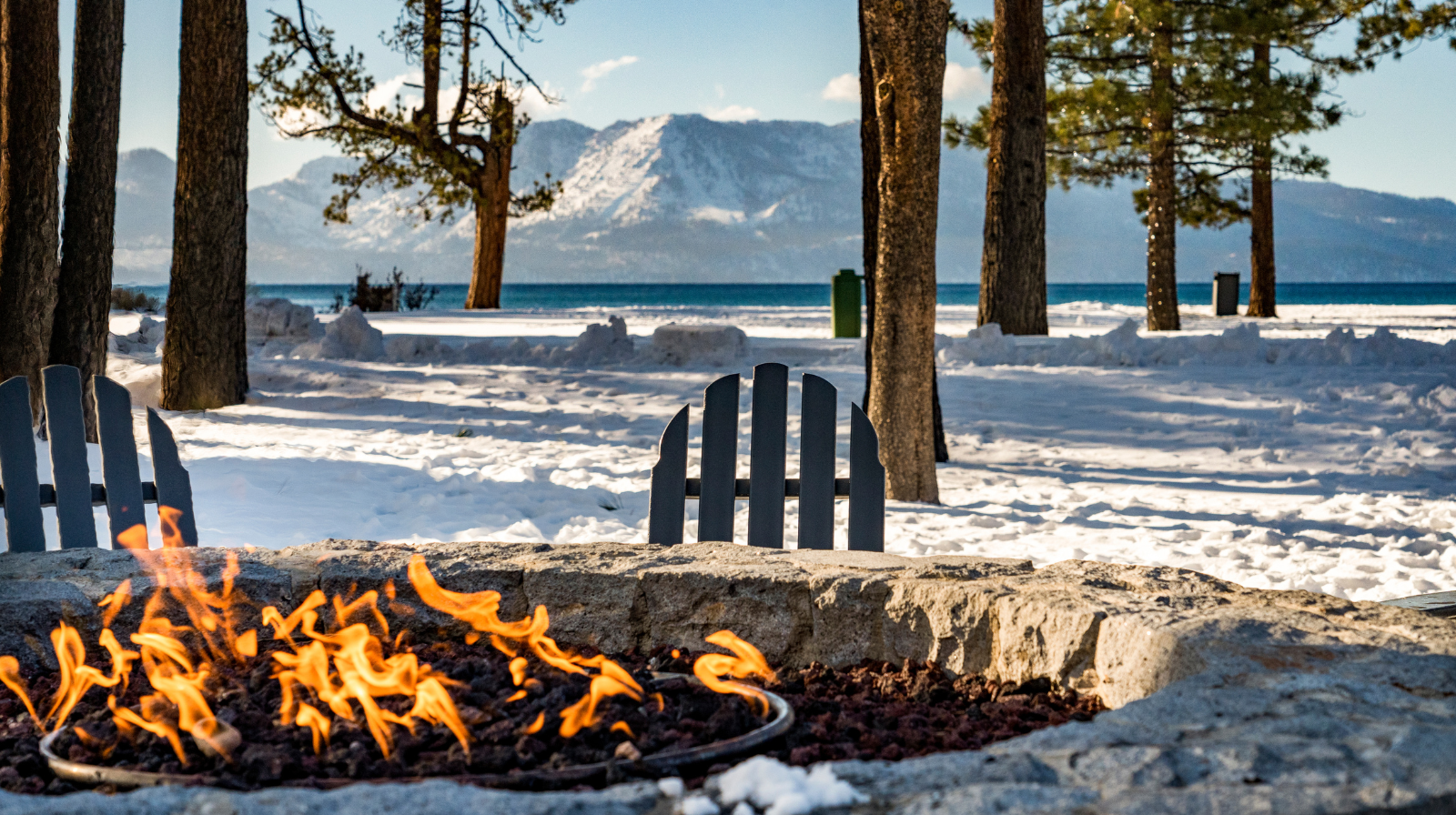 Winter fire pit on Lake Tahoe shoreline with snow-covered mountains and Adirondack chairs California Nevada