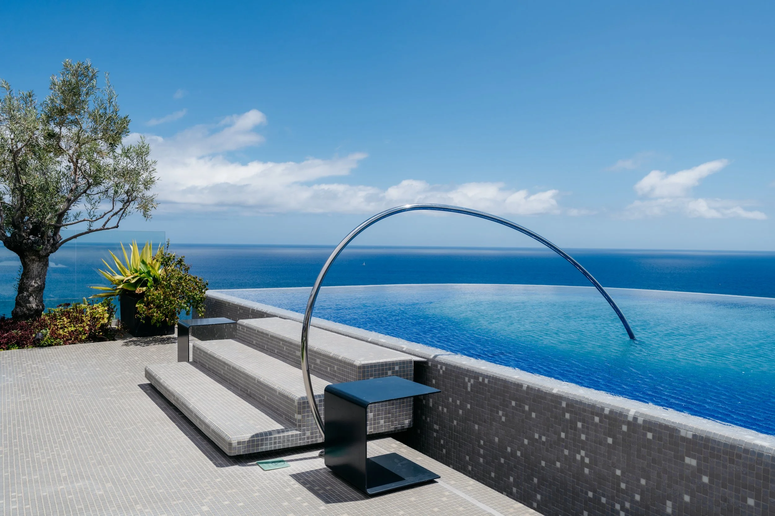 Modern infinity pool with sleek tile steps, sculptural metal arch, and unobstructed Atlantic Ocean views—contemporary design and tranquil setting in Funchal, Madeira.