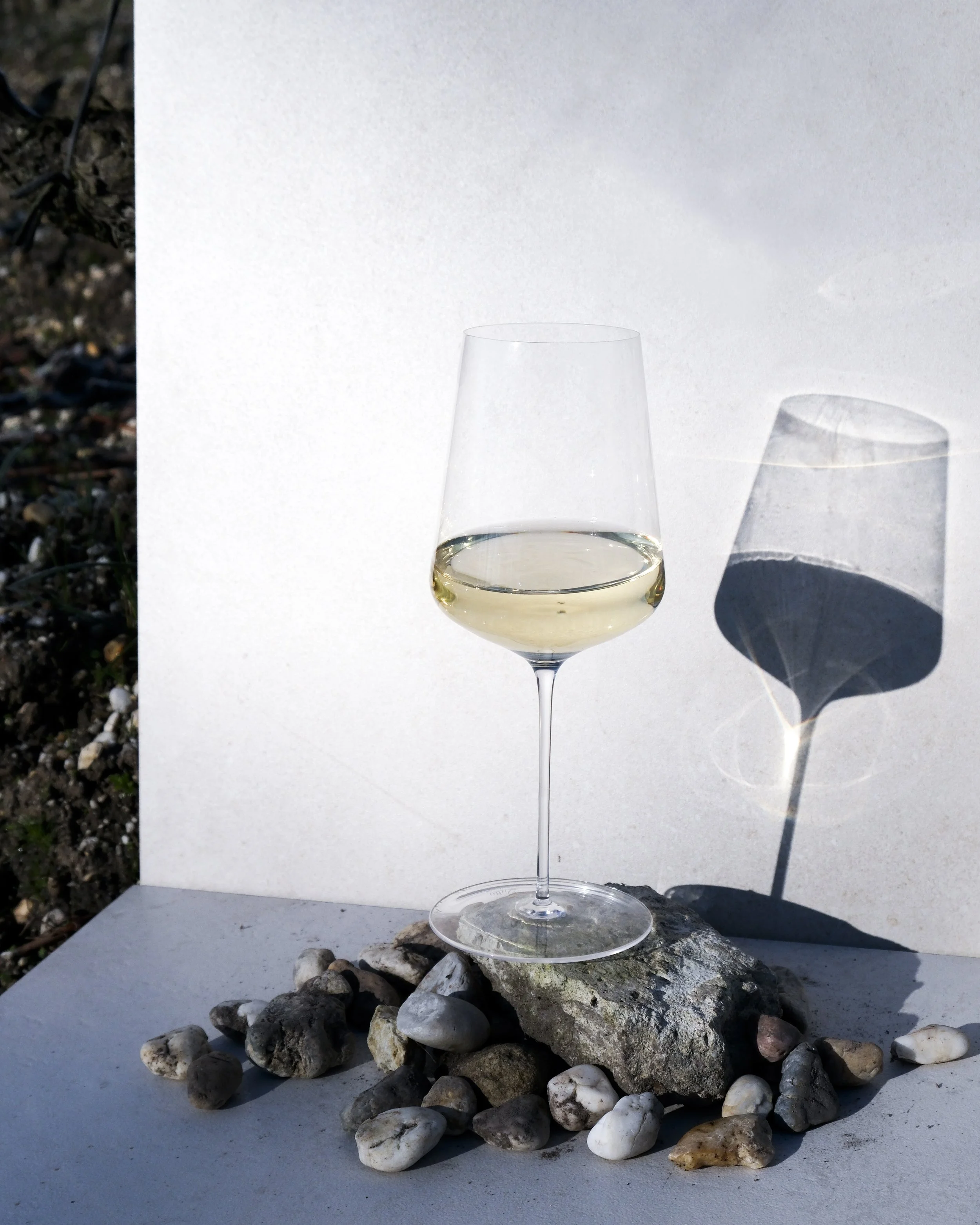 Verre de vin blanc sur un rocher entouré de petits cailloux, avec un fond blanc et une ombre du verre projetée sur le mur.