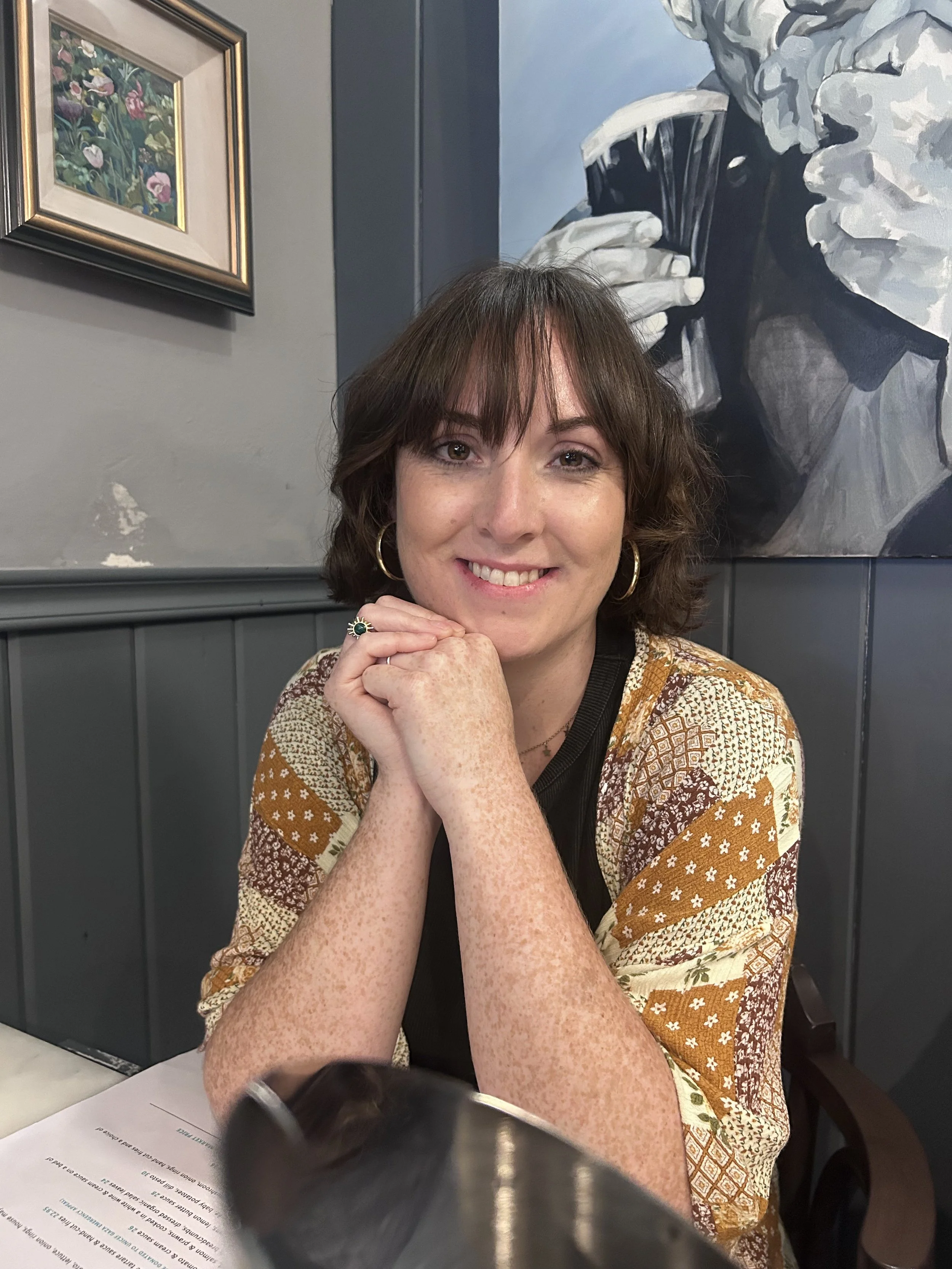 A woman with short brown hair smiling, resting her chin on her clasped hands, wearing gold hoop earrings and a patterned kimono-style top sitting at a table in a restaurant.