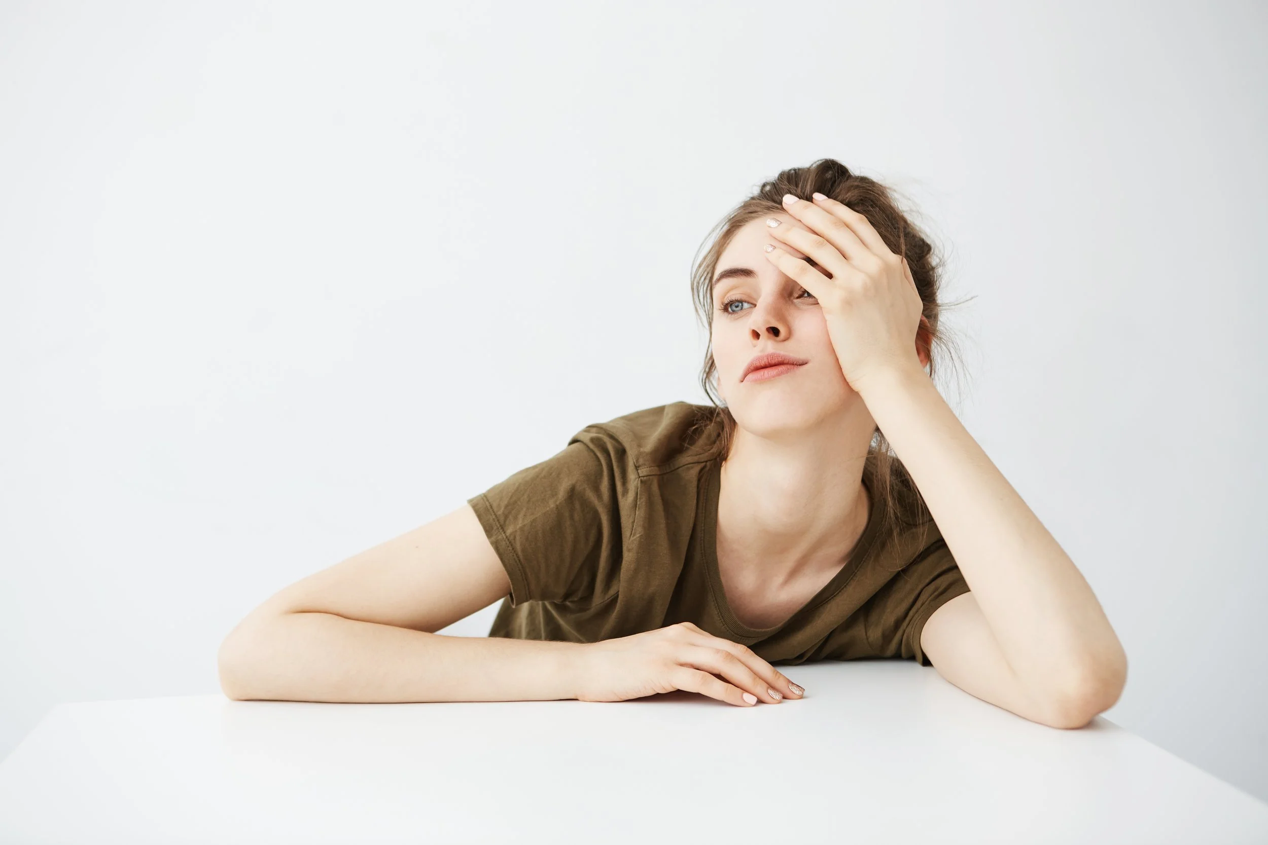Young woman with brown hair wearing an olive-green t-shirt, resting her head on her left hand with a tired expression, sitting at a white table against a plain white wall.