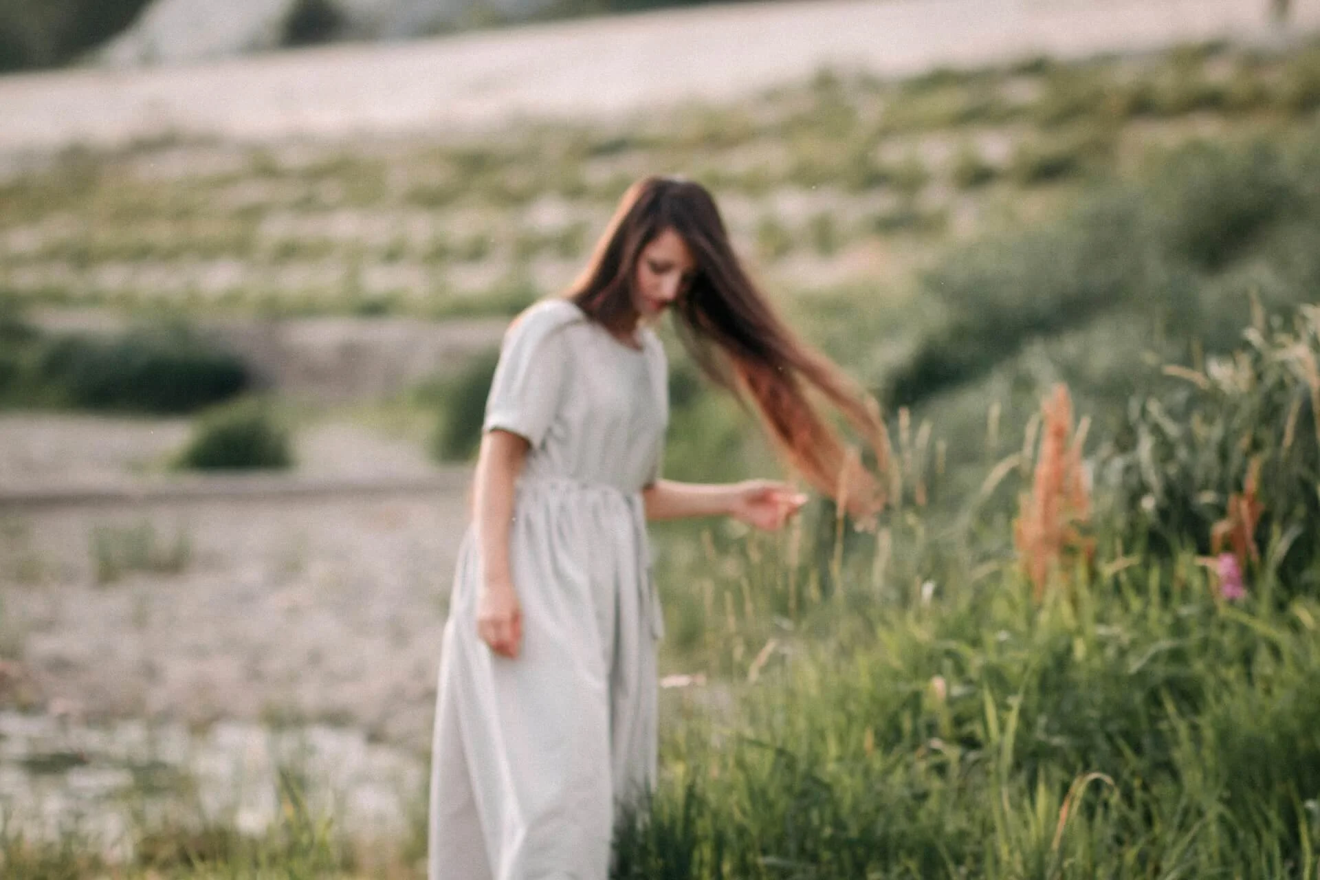 A woman in a long, light-colored dress standing outdoors in a green field, looking down at the grass.
