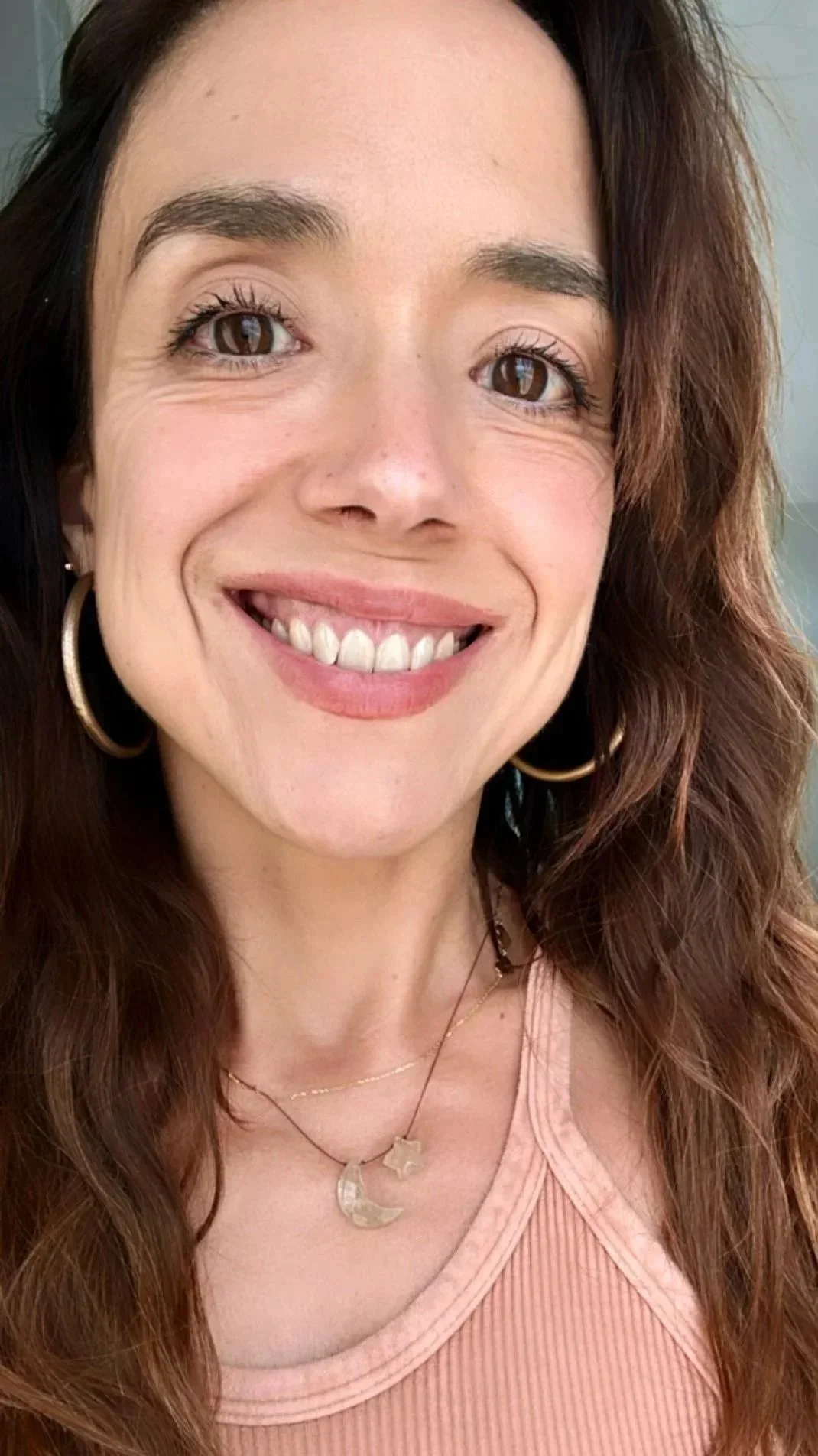 Close-up of a smiling woman with long wavy auburn hair, brown eyes, wearing hoop earrings and layered necklaces, dressed in a pink ribbed tank top.