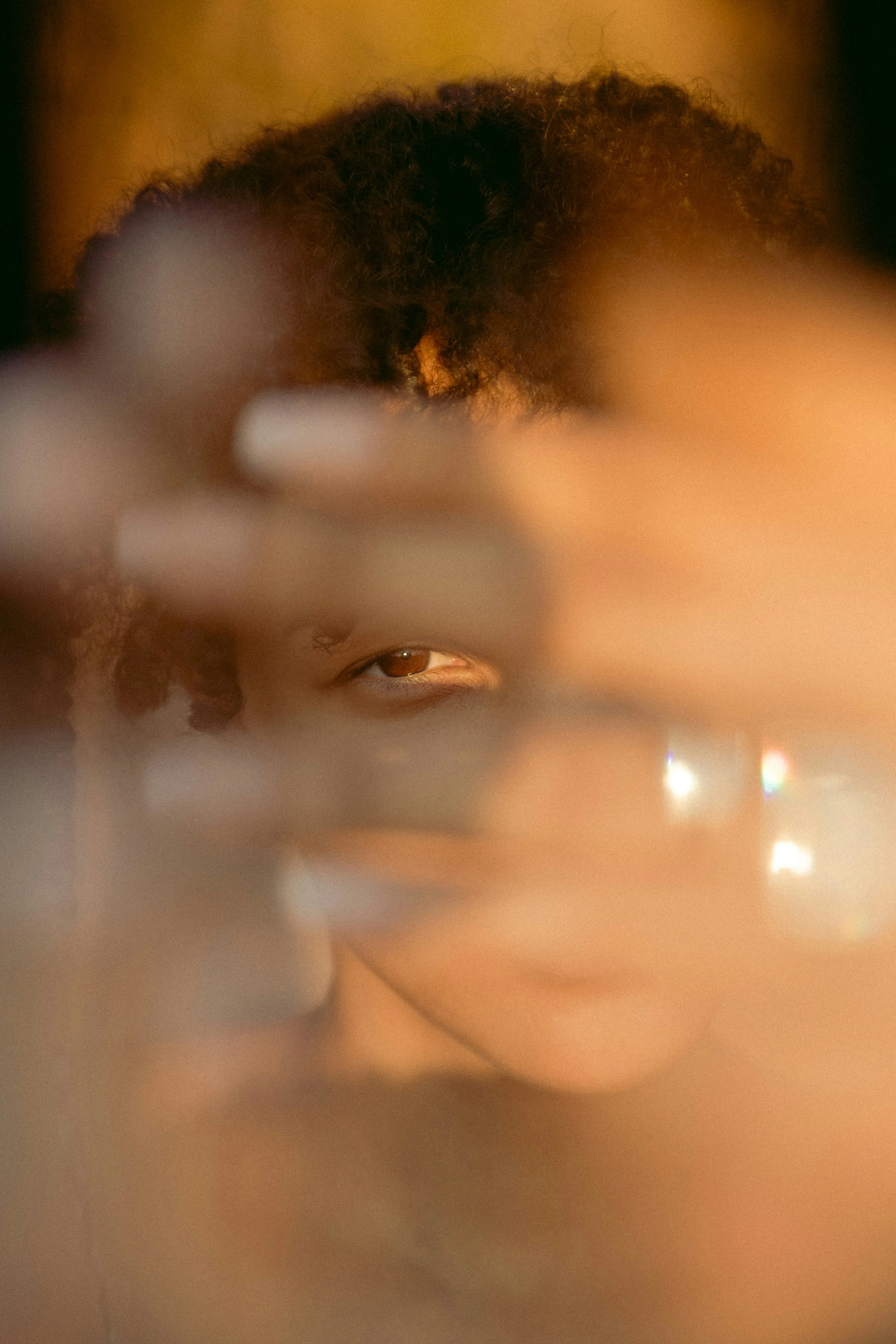 Close-up of a person with curly hair, partially obscured by out-of-focus hand and glasses, with one eye visible through a gap, warm lighting