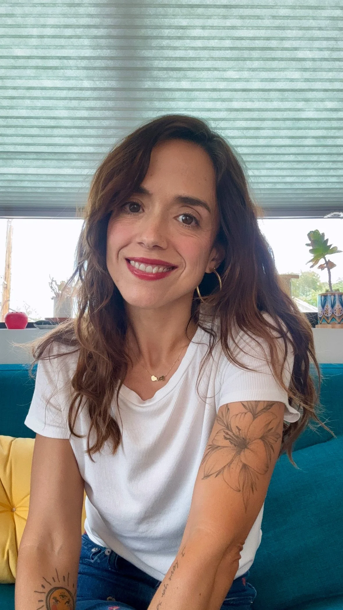 A woman with wavy brown hair, smiling, wearing a white T-shirt, sitting on a colorful couch with yellow and blue cushions, and displaying tattoos on her arms. There are plants and decorative items on the window sill behind her.