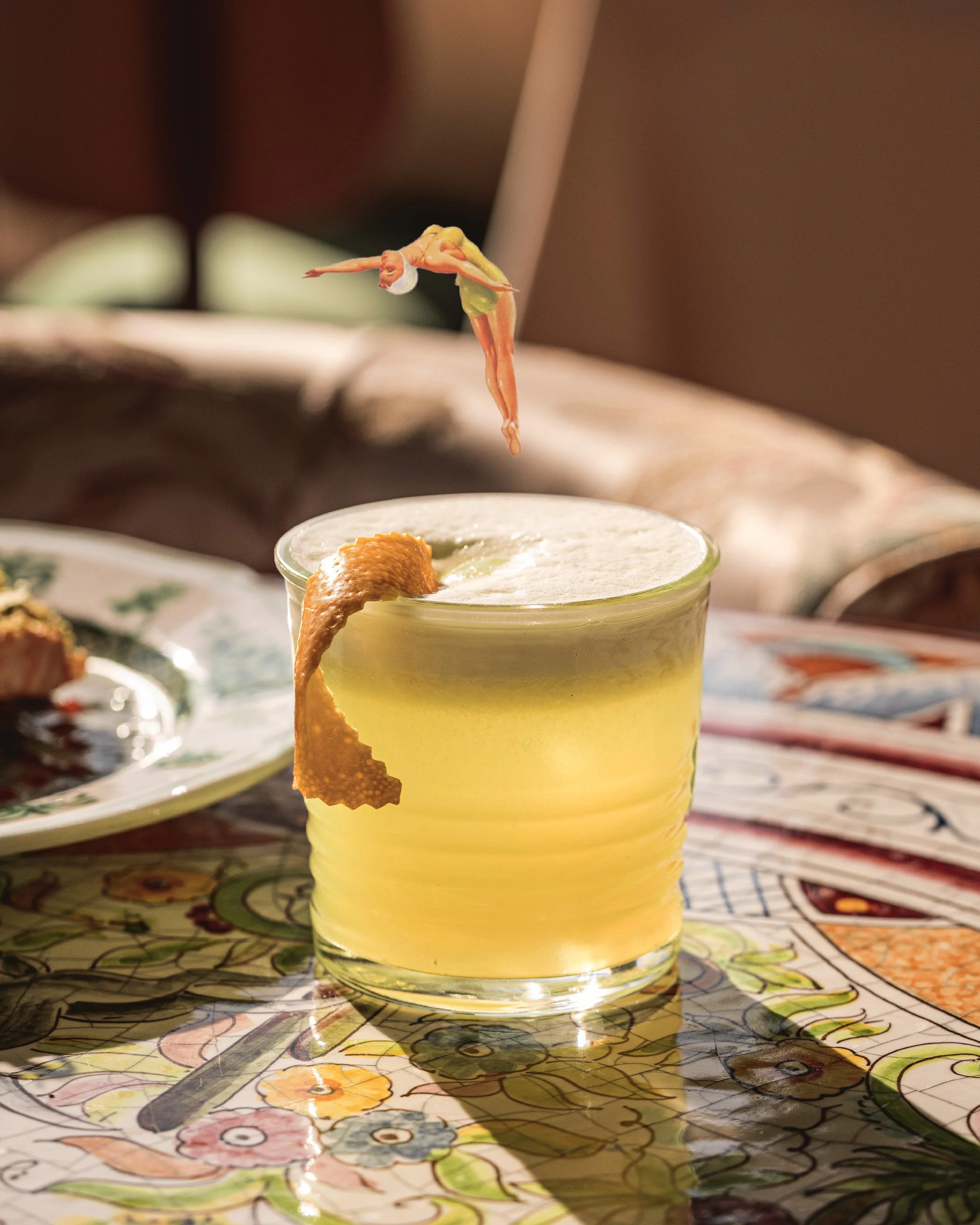 A yellow cocktail with lemon peel garnishment on a colorful table, with a small figure of a person in vacation attire floating above it.