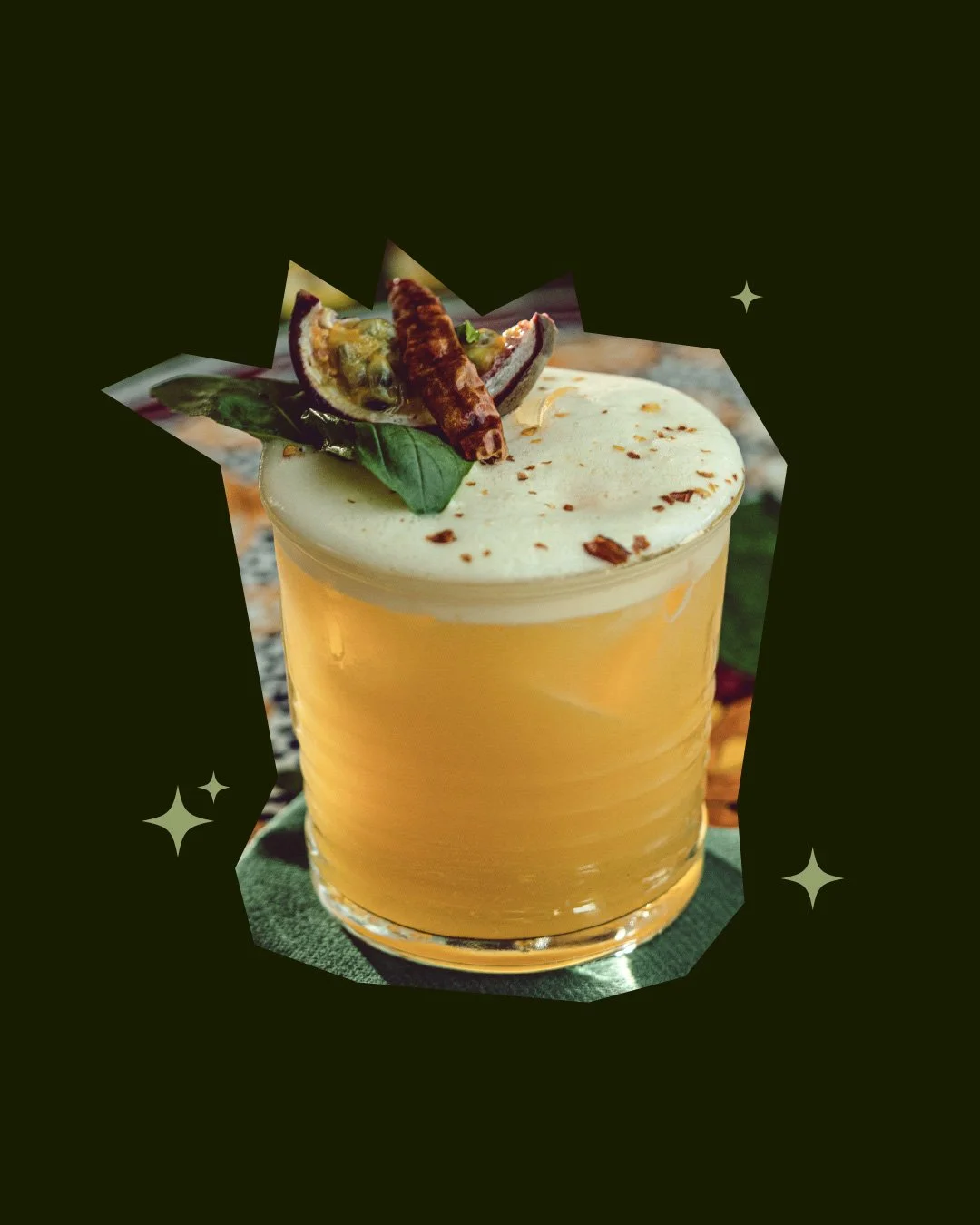 A yellow cocktail in a clear glass topped with whipped cream, chocolate shavings, a basil leaf, and a tamarind drink with a chili pepper