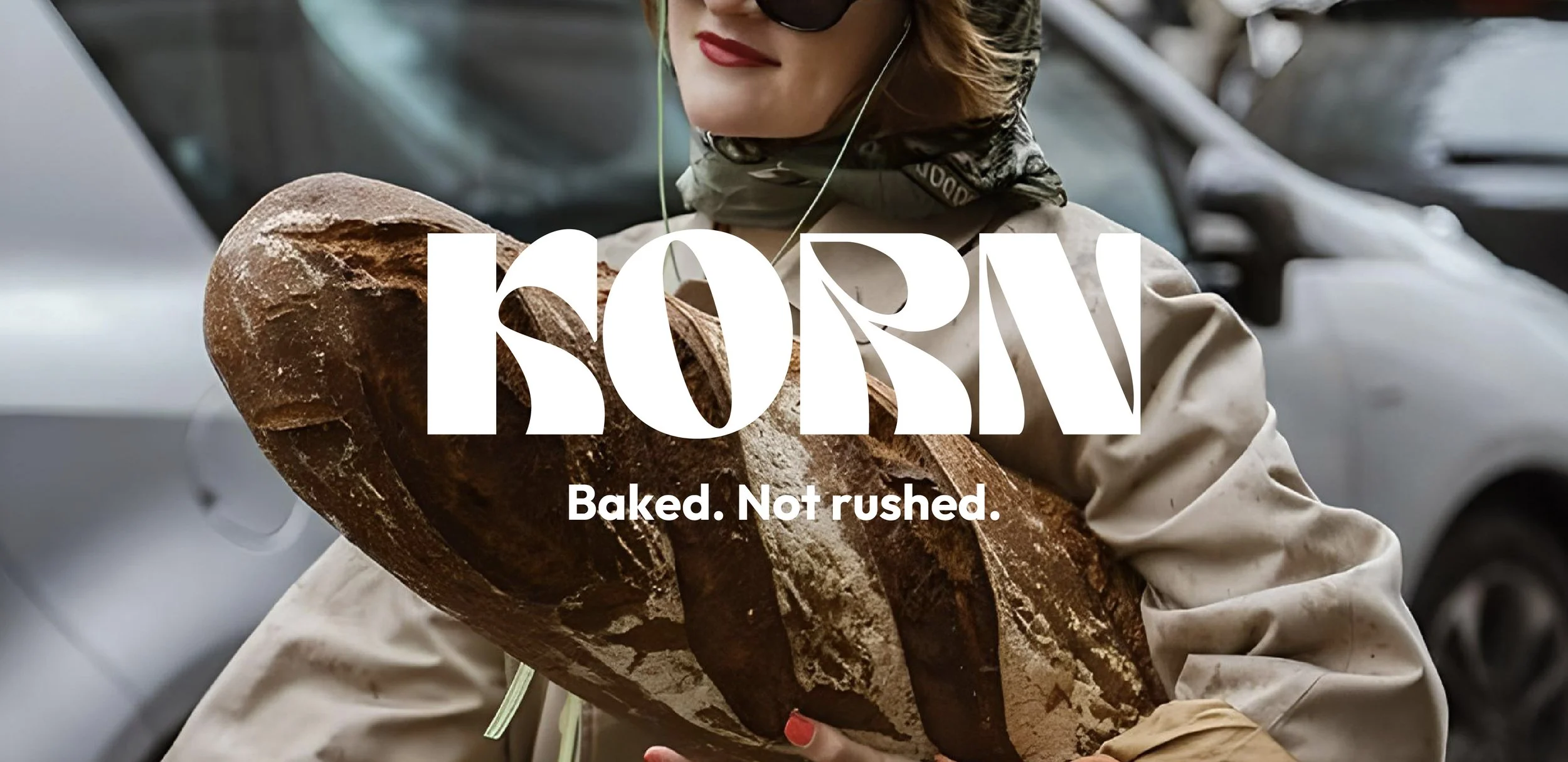 Person holding a large loaf of freshly baked bread outdoors, wearing sunglasses, a beige coat, and a green scarf with the text "KORN" and "Baked. Not rushed." overlaid on the image.