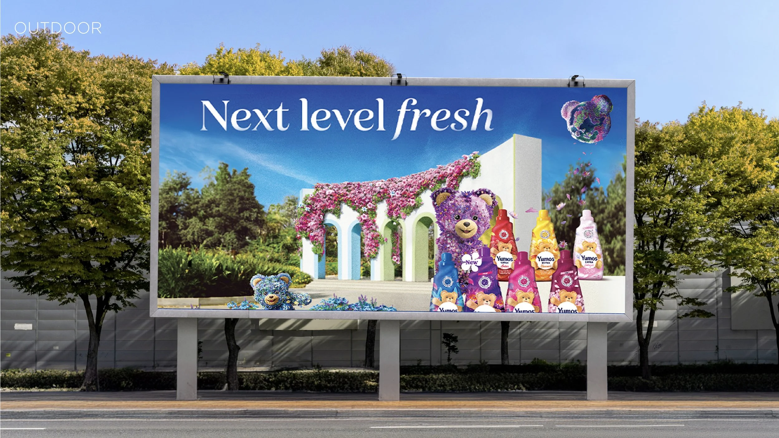 Outdoor billboard advertising Yumo's bear-themed laundry detergent bottles in a colorful garden scene with a white building covered in pink flowers, and a large purple bear mascot among smaller bear plush toys. The text reads 'Next level fresh'.