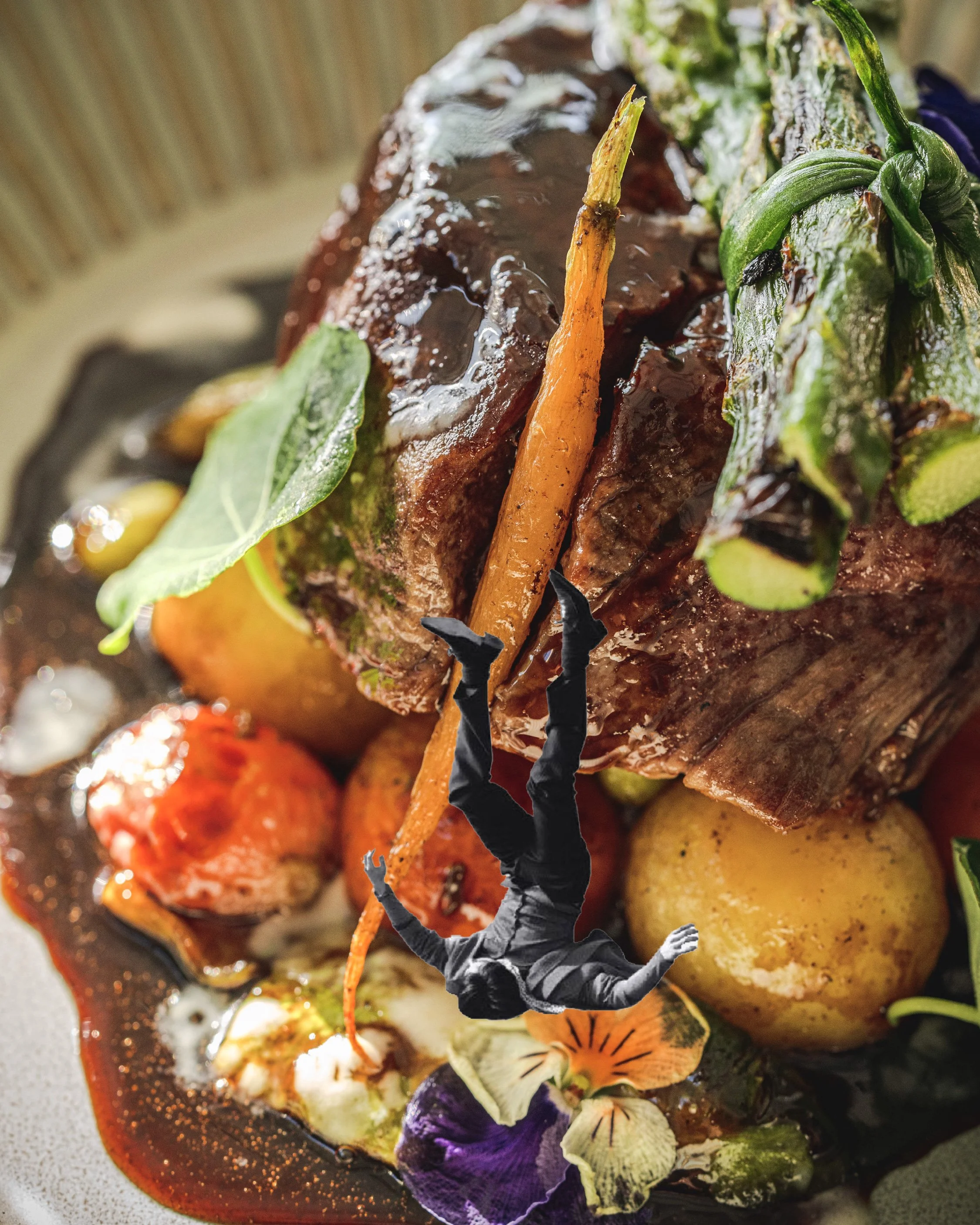 Close-up of a gourmet meat dish garnished with herbs, vegetables, and edible flowers, with a miniature person hanging upside down from a carrot.