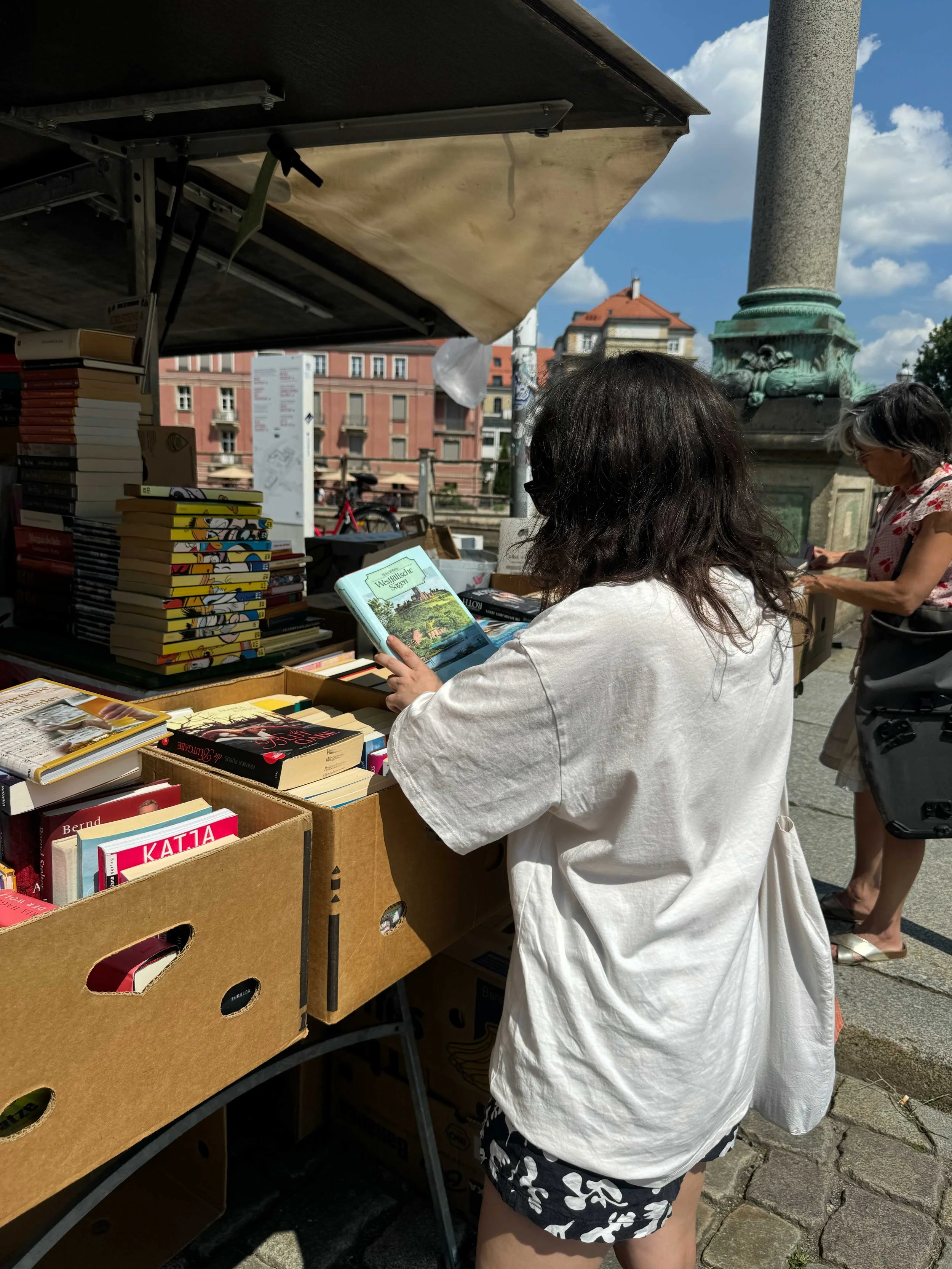 Woman browsing books at an outdoor street bookstore with other shoppers nearby, under a canopy on a sunny day.