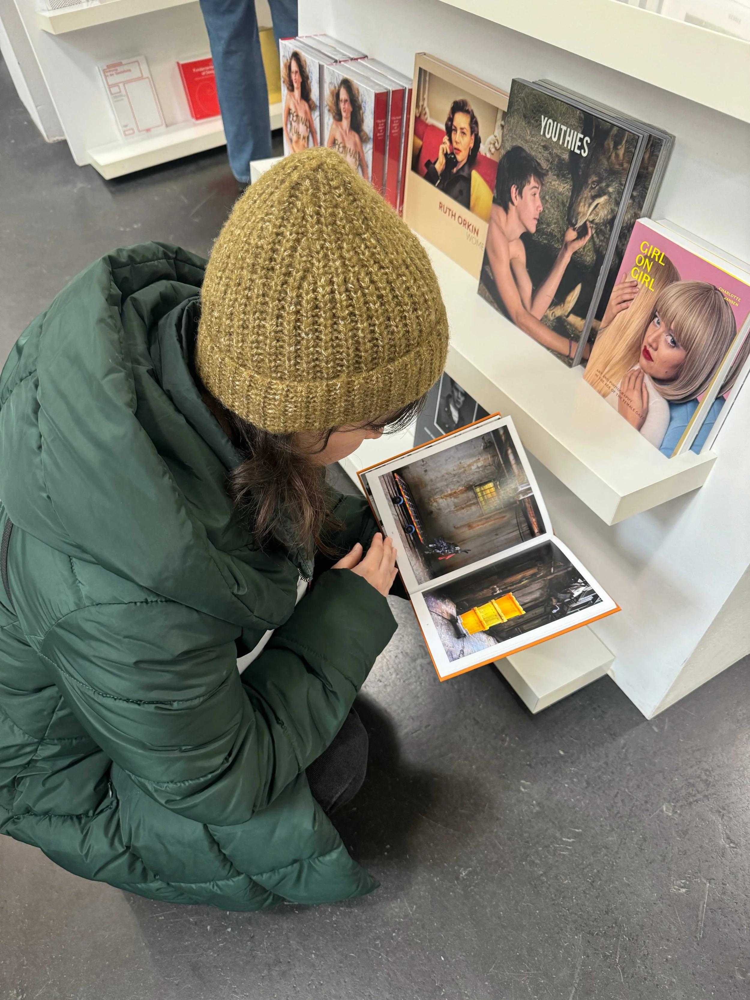 A woman wearing a green jacket and a brown knit hat is sitting on the floor, looking at a photo book in a bookstore or gallery. There are shelves with fashion and art books behind her, with titles like 'YOUTHIES', 'RUTH ORKIN WOMEN', and 'GIRL ON GIR