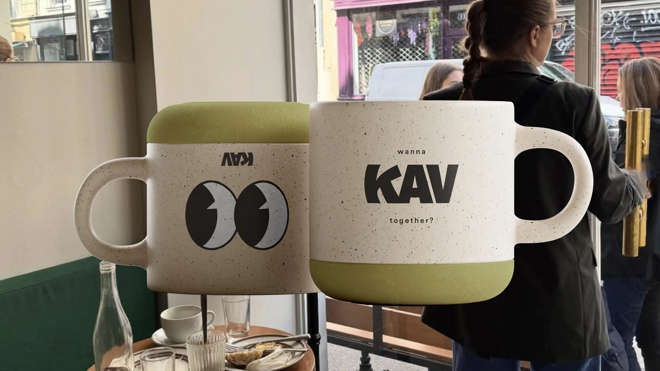 Two ceramic mugs with speech bubble graphics placed in a creative display, with people standing near a window in a cafe or restaurant setting.