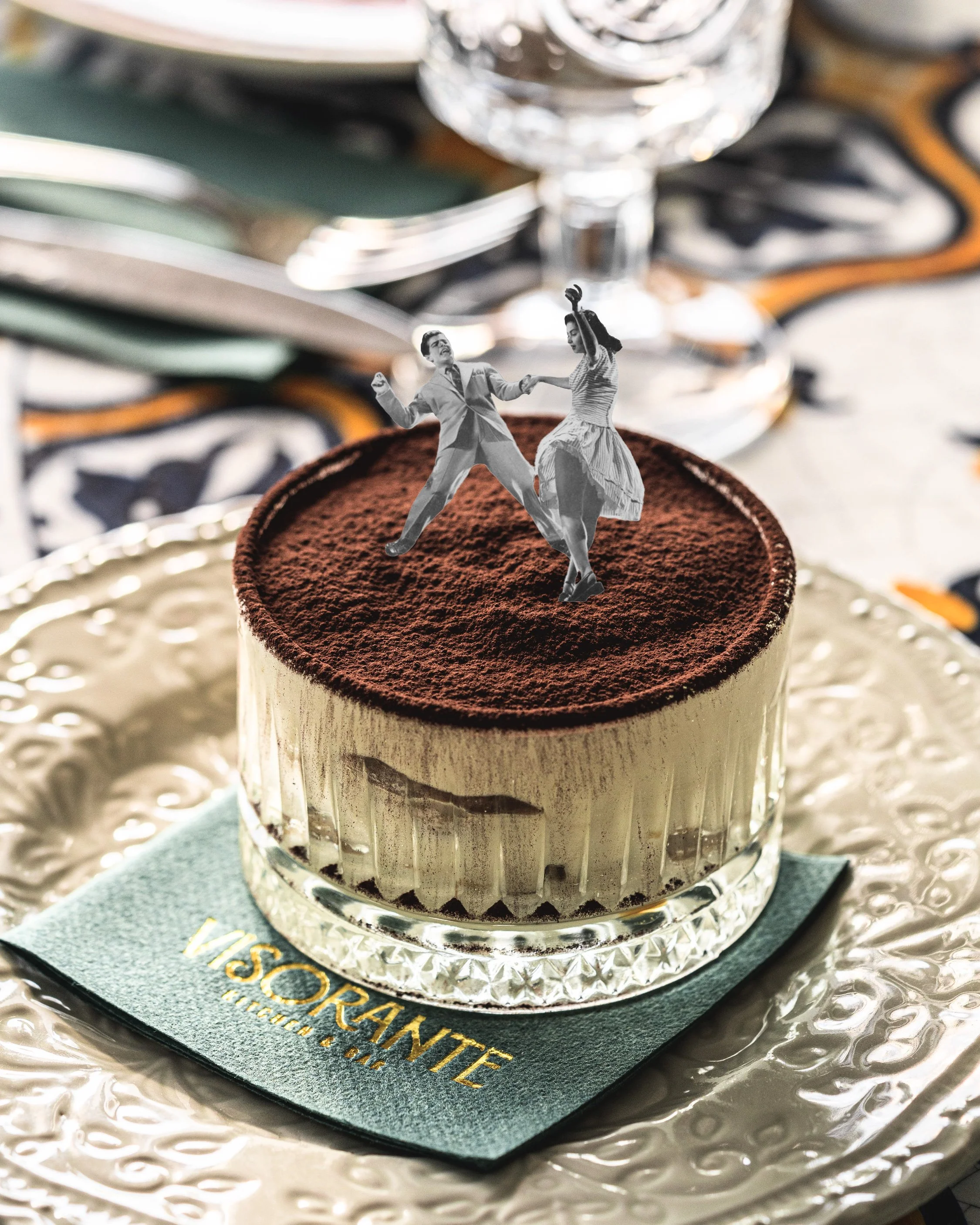 A dessert topped with cocoa powder featuring a paper cutout of a dancing couple, placed on a decorative plate with a green napkin underneath.