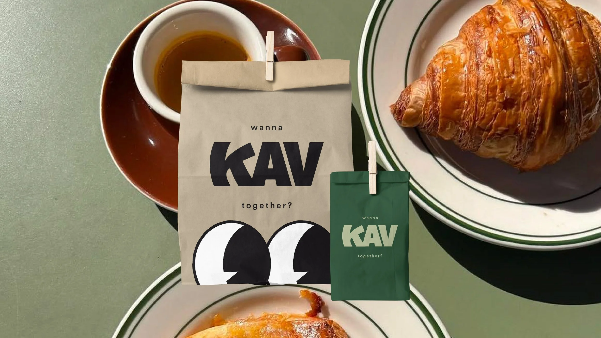 A cup of coffee on a saucer, a croissant on a plate, and two paper bags with the words 'wanna KAV together?' printed on them, set on a green surface.