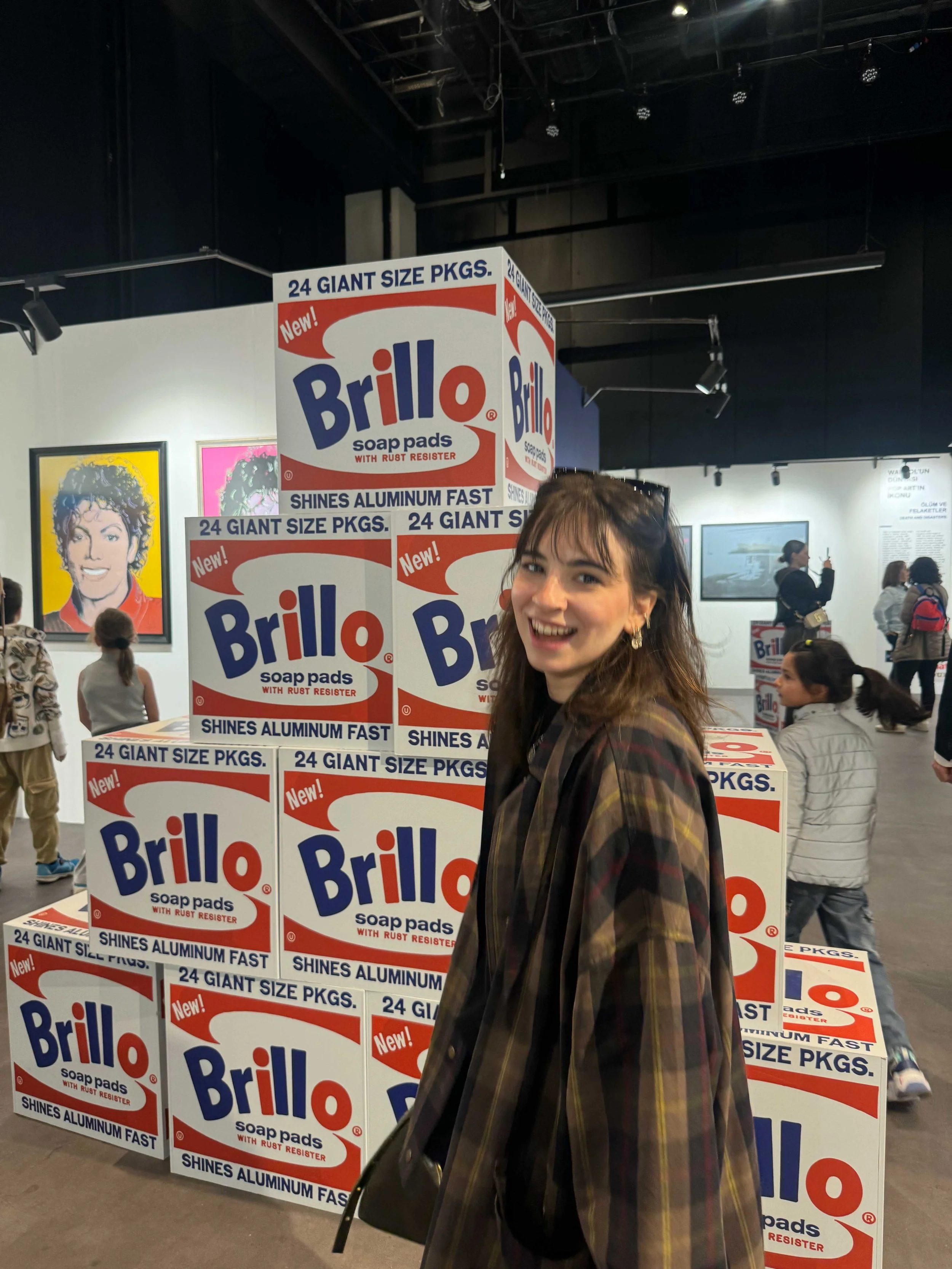 A smiling woman with dark hair, wearing a plaid jacket, standing in front of stacked boxes of Brillo soap pads in an indoor setting, with other people and artwork in the background.