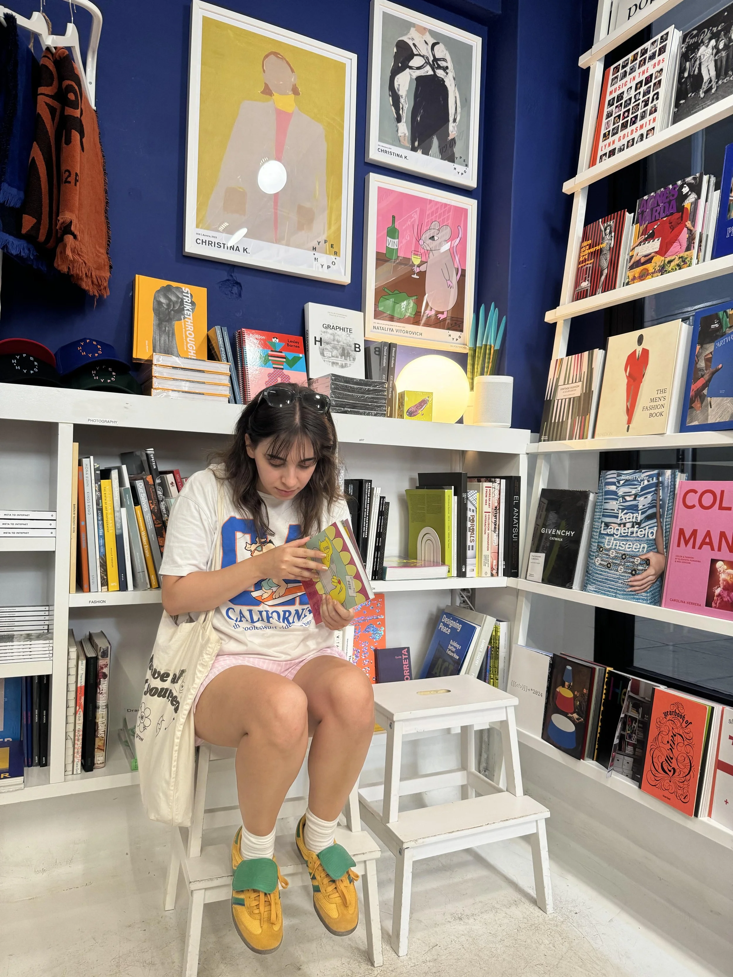 A young woman sits on a white stool in a bookstore, flipping through a colorful book. The background features shelves filled with books, artwork on the blue walls, and various decorative items.