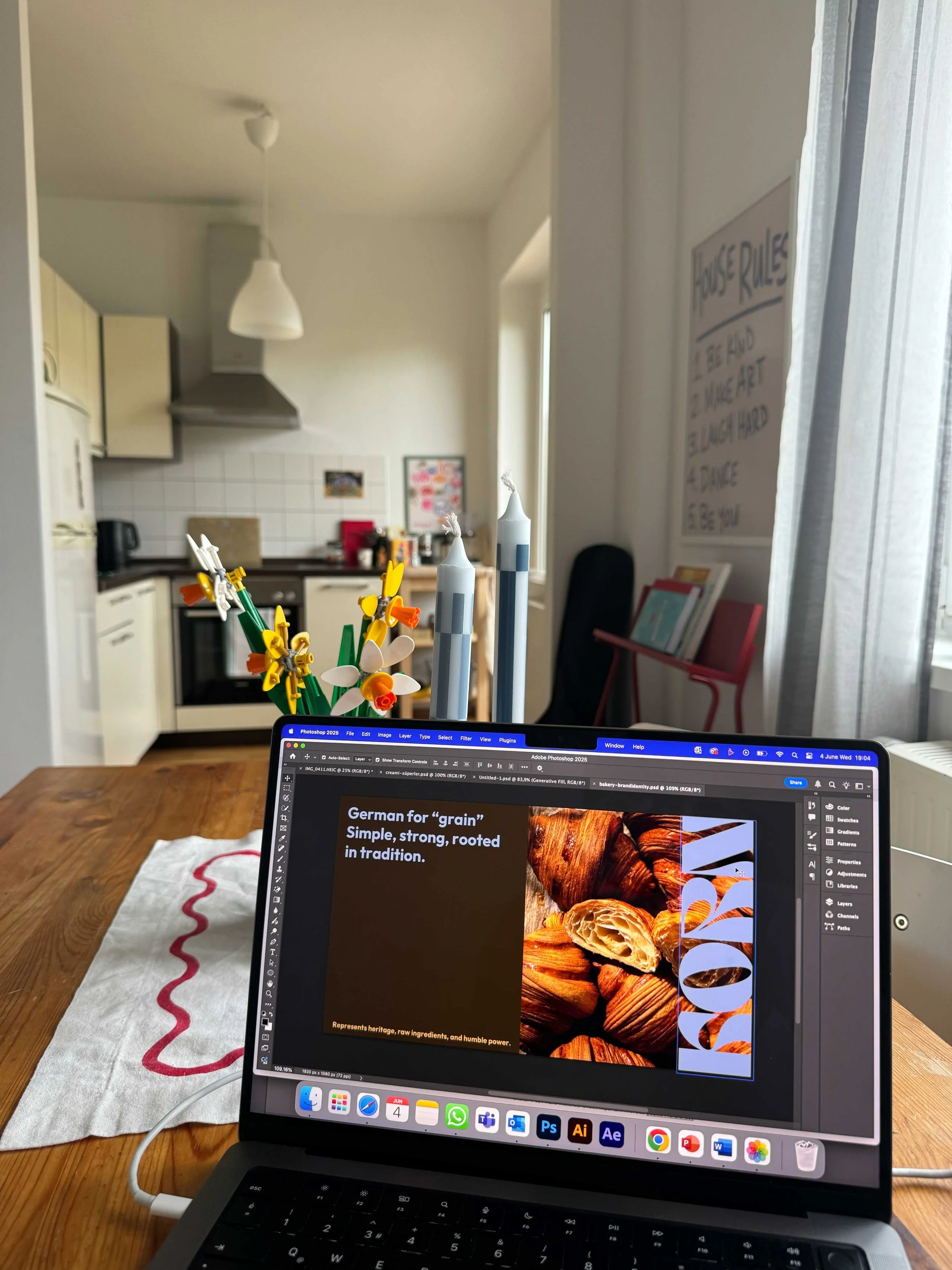 Inside a kitchen and dining area, a laptop displays a presentation about German bread, with a foreground of lit candles and a vase with yellow and white flowers on the dining table.