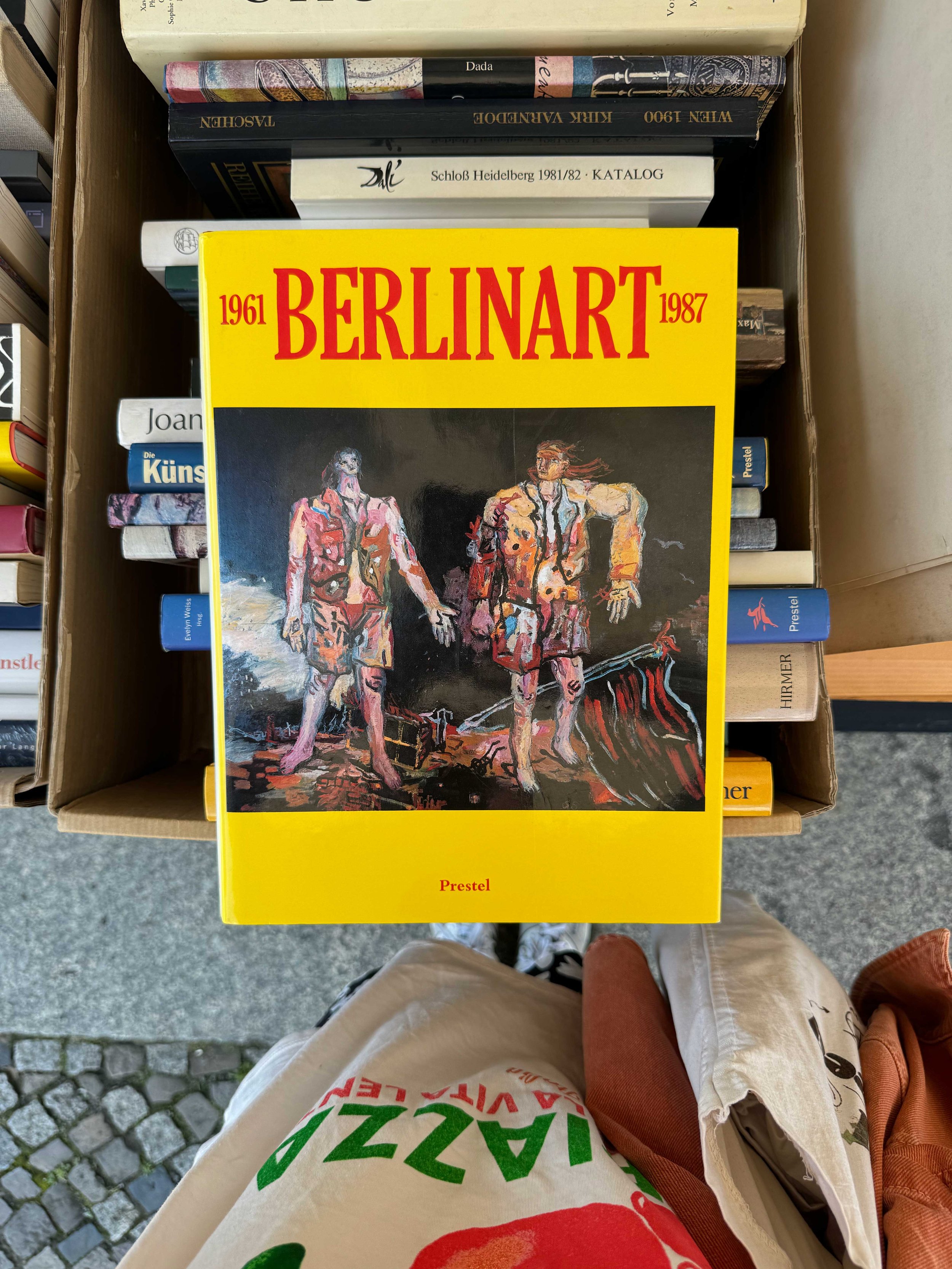 A book titled 'BERLINART 1961-1987' with a yellow cover, featuring an abstract painting of two figures standing on a rocky terrain, in front of a dark background.