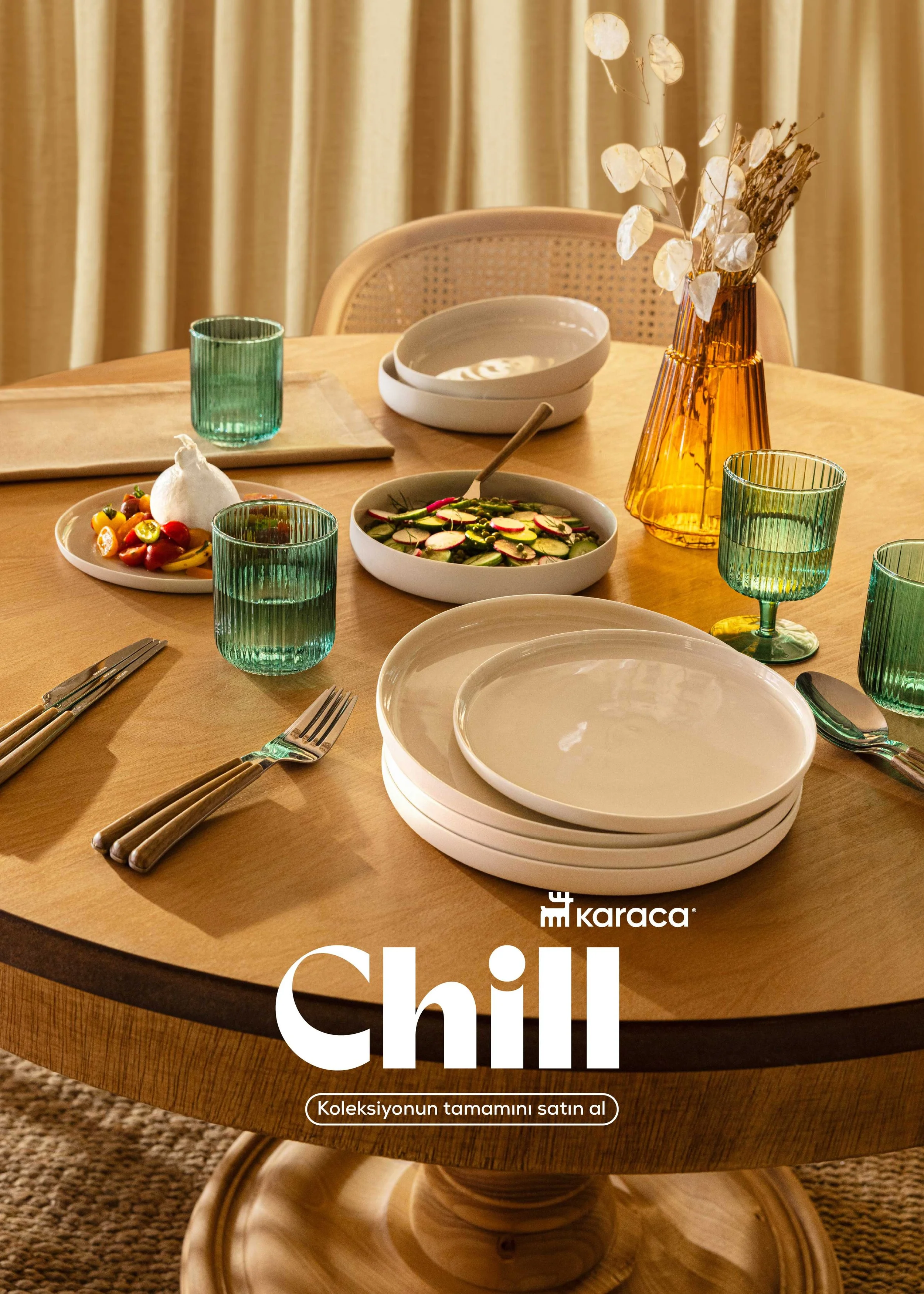 A round wooden dining table set with white plates, green drinking glasses, silverware, and bowls of salad. There is a tall orange vase with dried white leaves and a smaller white bowl with a round white item. The background features beige curtains.