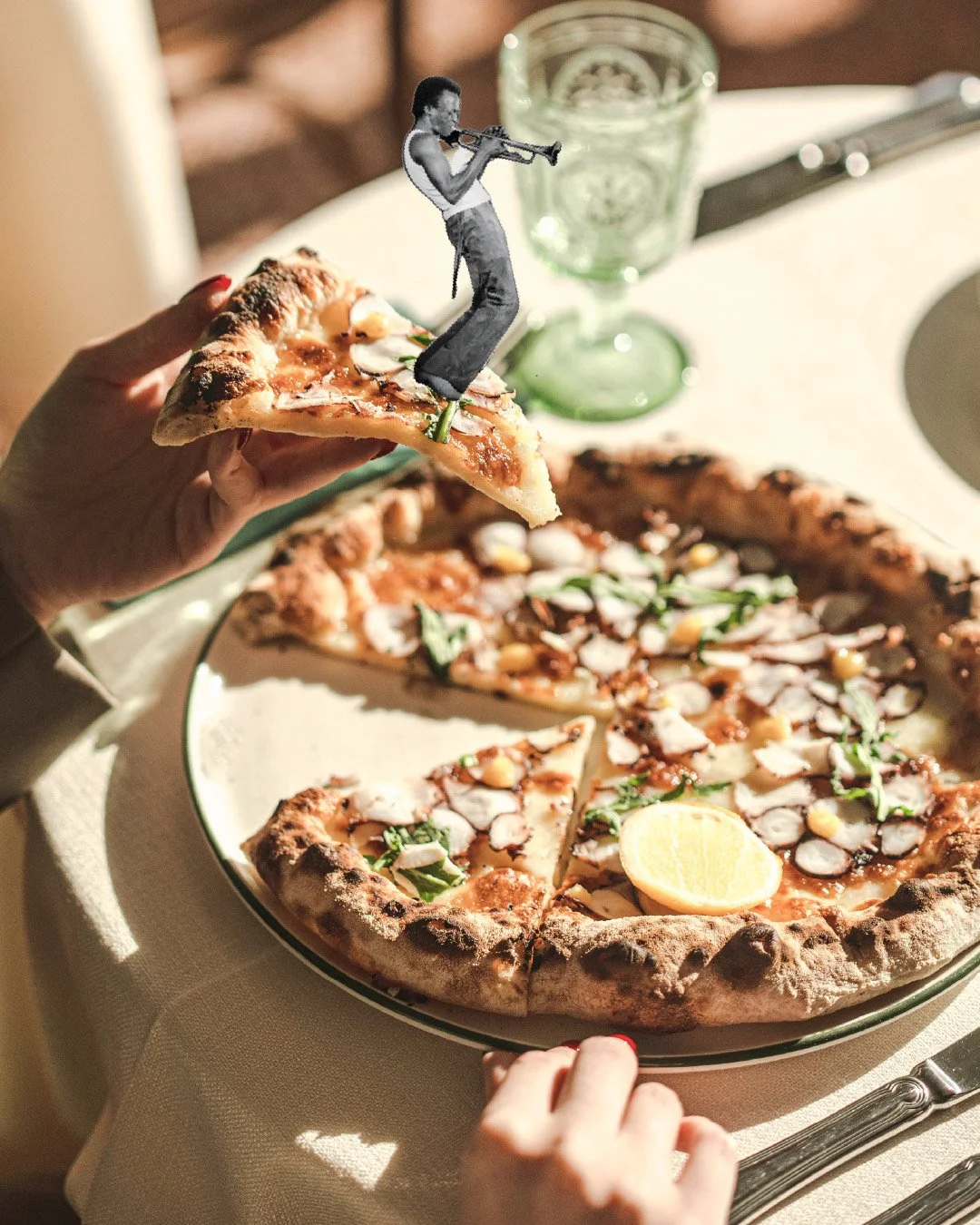 A plate of pizza with a lemon wedge and toppings, on a table with a glass of water and a knife in the background; a person is holding a slice with a small illustration of a man in a suit playing a trumpet sitting on the slice.