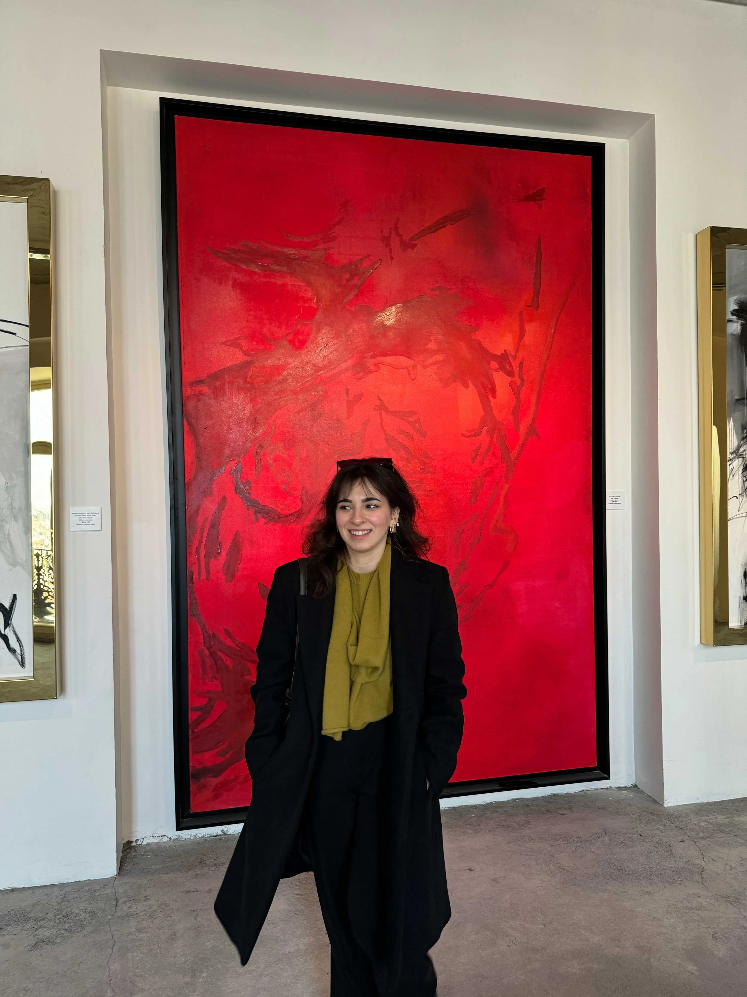 A woman with dark hair and a black coat standing in front of a large red abstract painting at an art gallery.