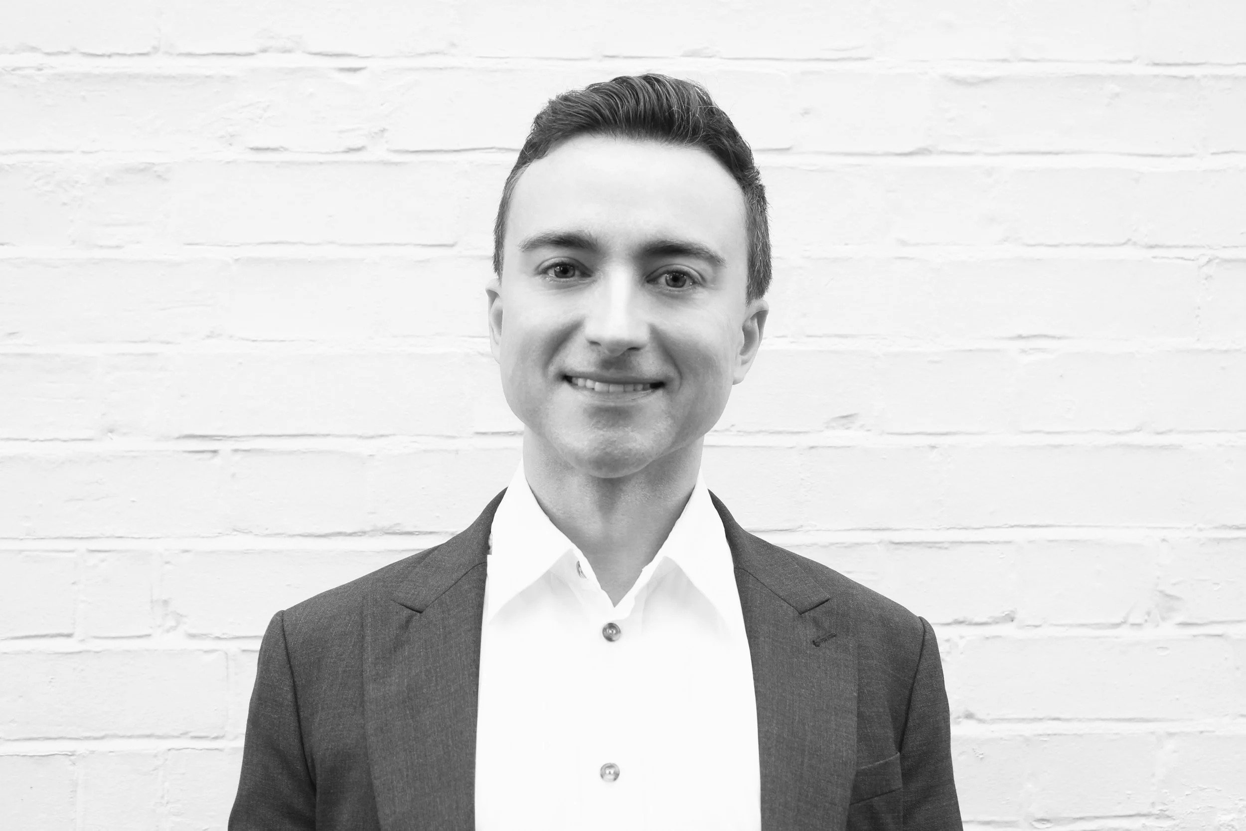 Black and white portrait of a smiling young man in a suit and white shirt, standing in front of a brick wall.