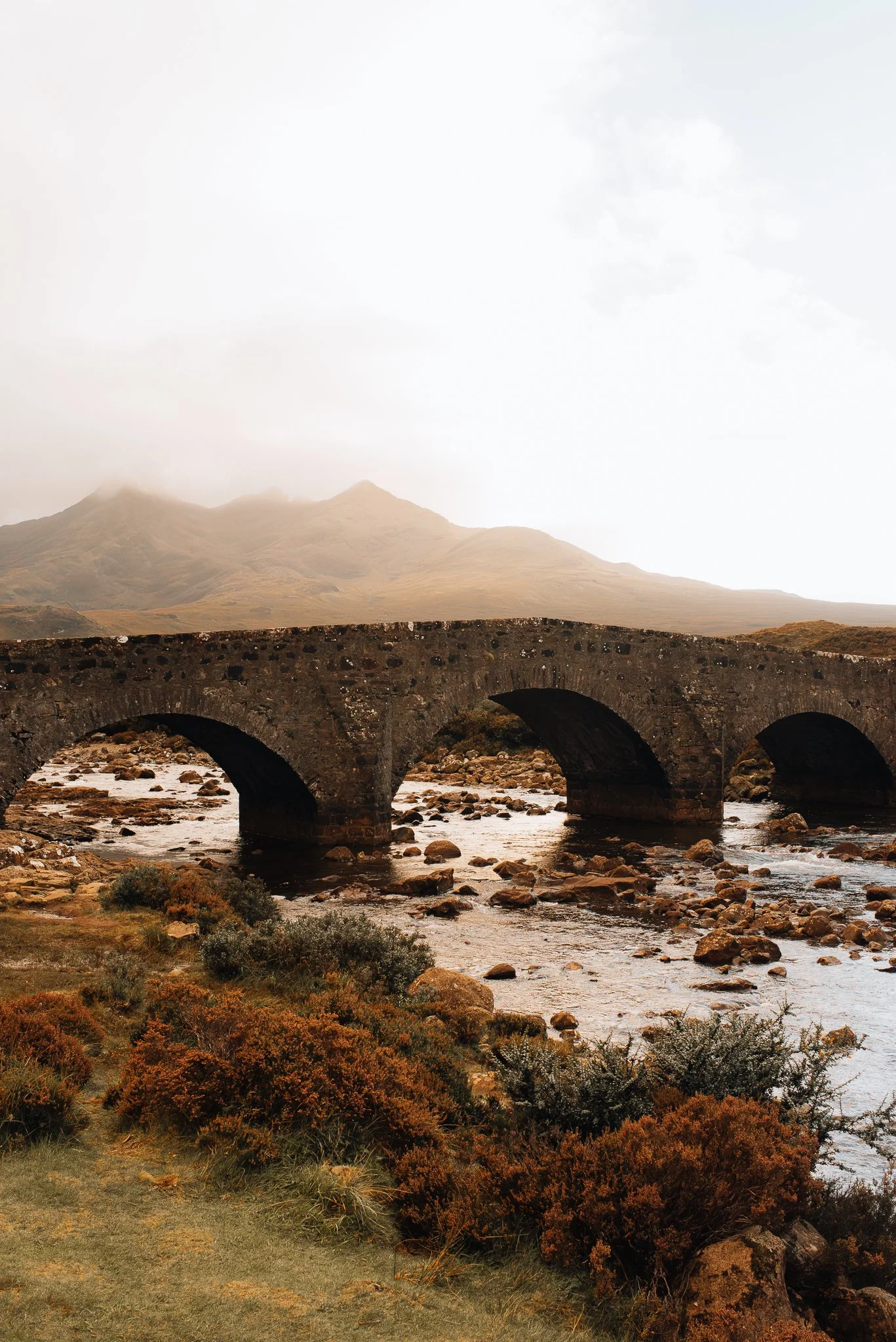 An old stone bridge over a river with rocks, mountains in the background, and cloudy sky.
