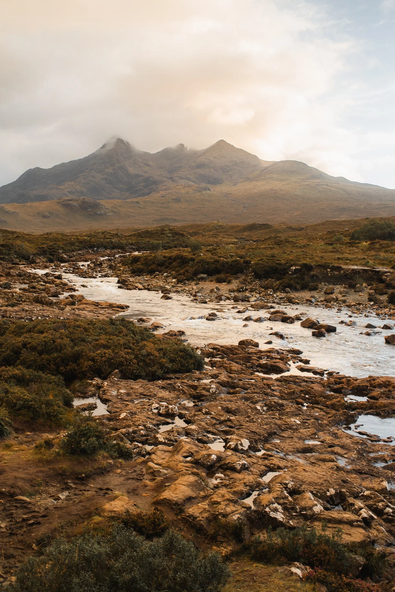 Scenic view of mountains with rocky river in the foreground, cloudy sky overhead.
