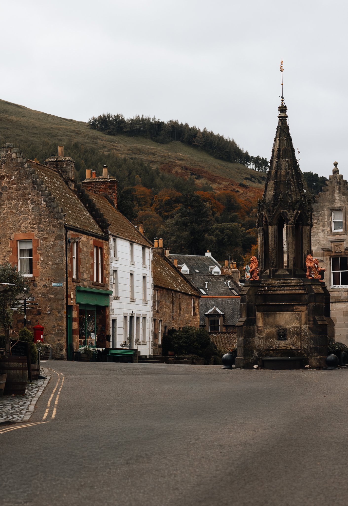A small town street with stone buildings, a monument, and rolling hills in the background during overcast weather.
