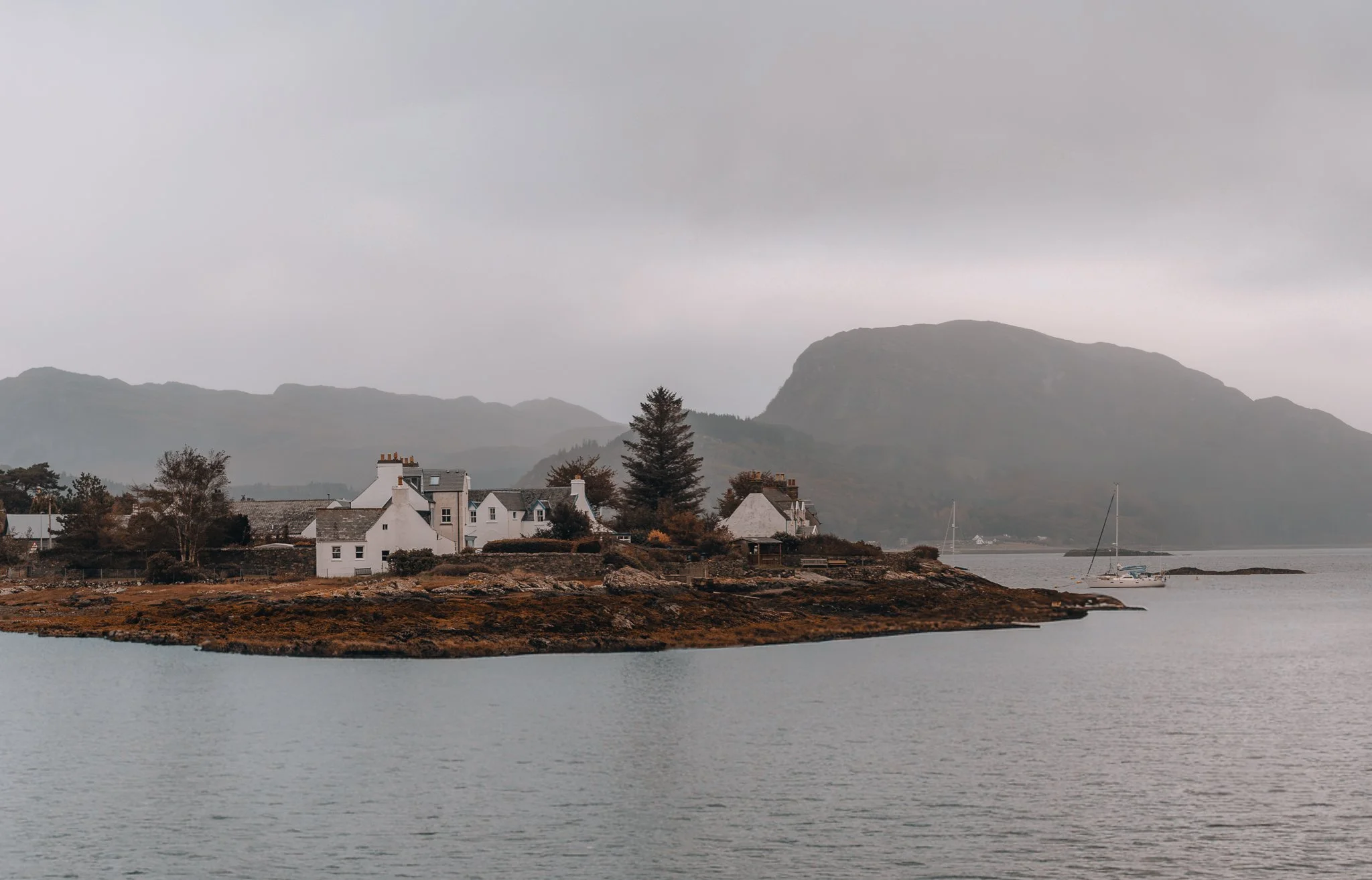 The small coastal village of Plockton with white houses and chimneys, surrounded by trees and mountains, next to a body of water with sailboats, under a cloudy sky.