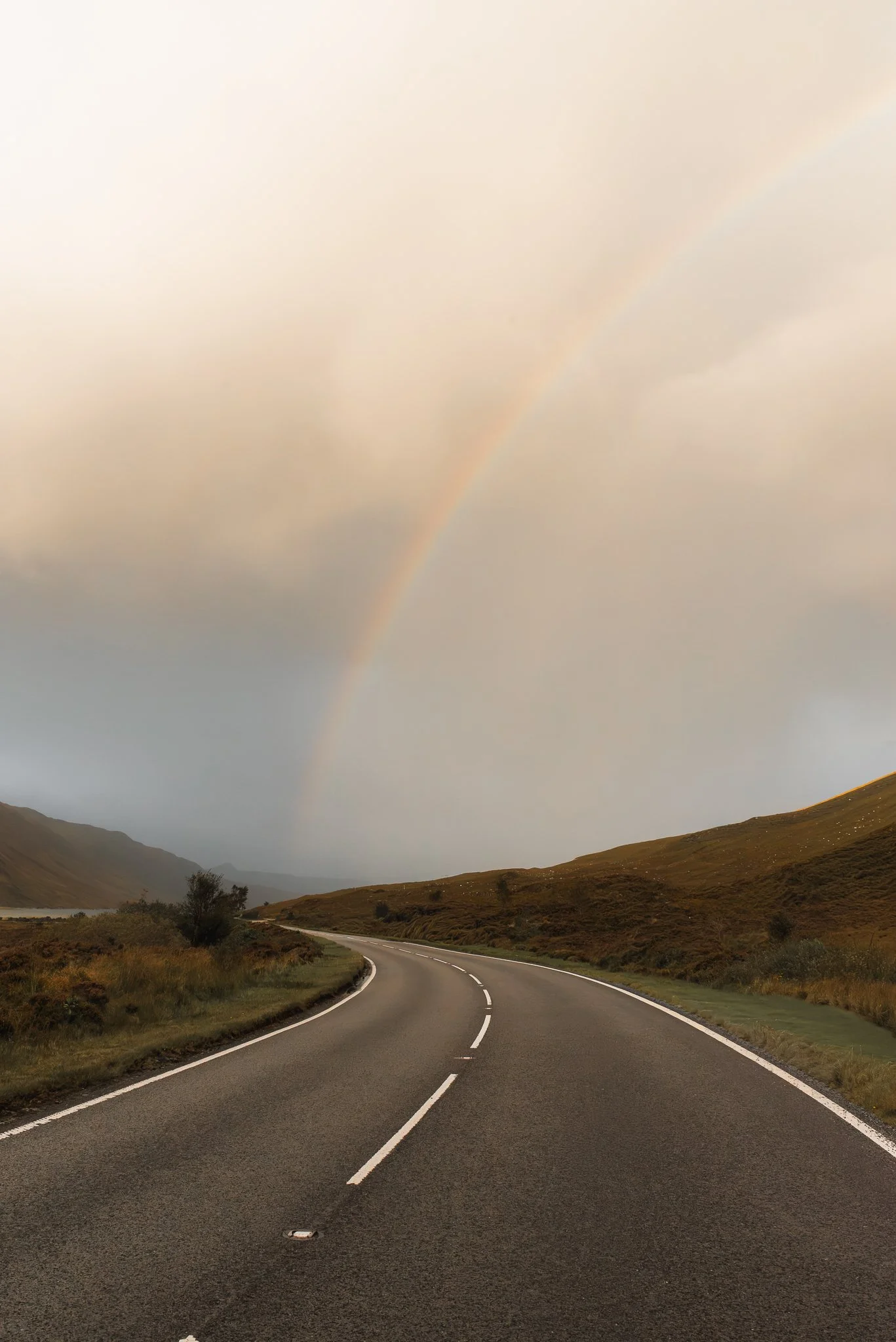A winding road runs through a hilly landscape with a faint rainbow in the cloudy sky.