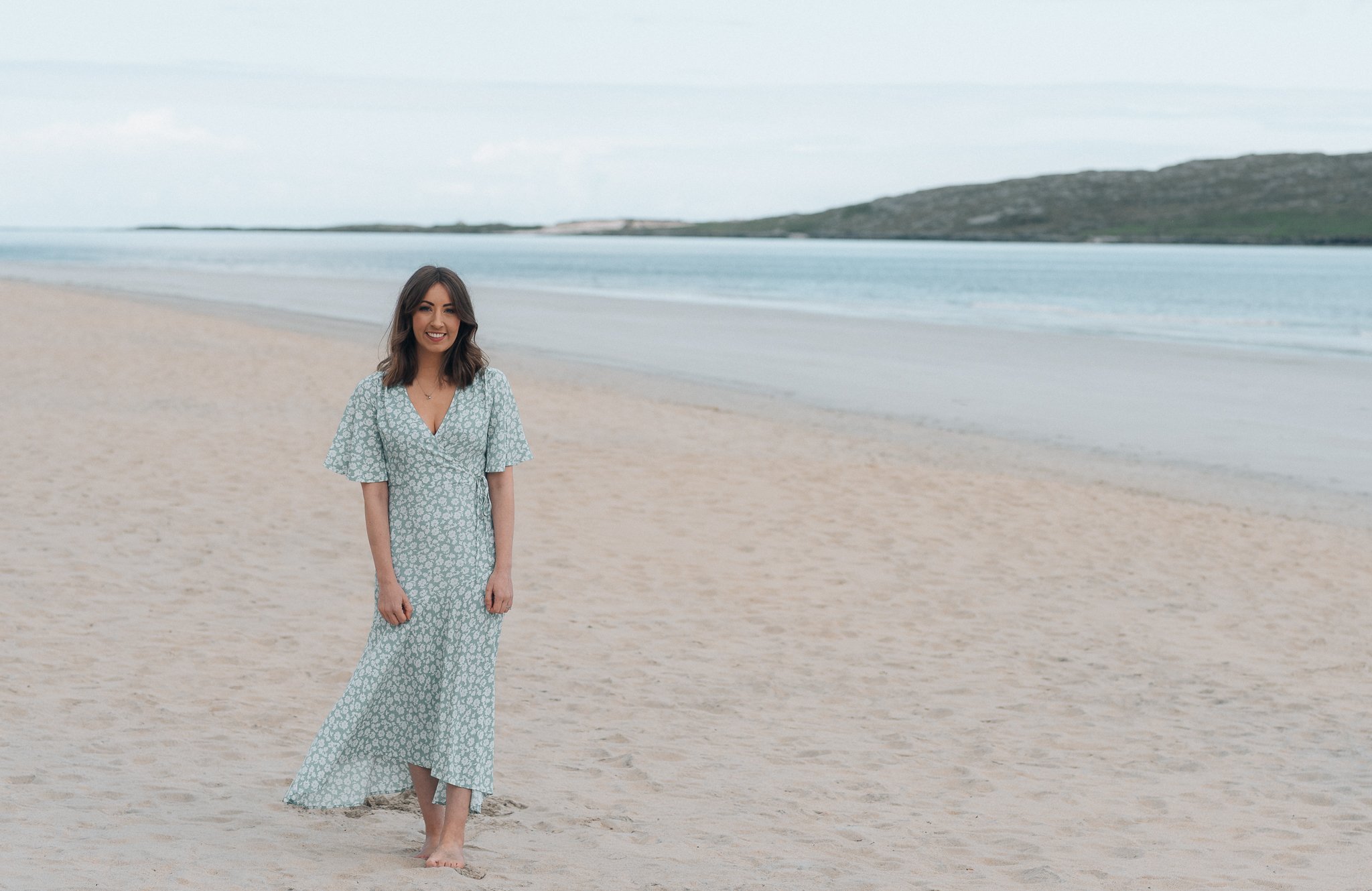 A woman in a patterned dress walking barefoot on the beach with the ocean and hills in the background.
