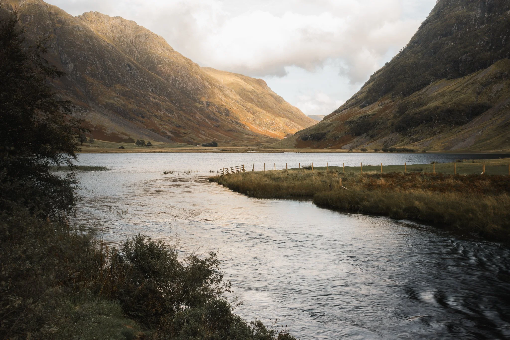 A river flowing through a valley surrounded by mountains with sparse vegetation, under a partly cloudy sky.